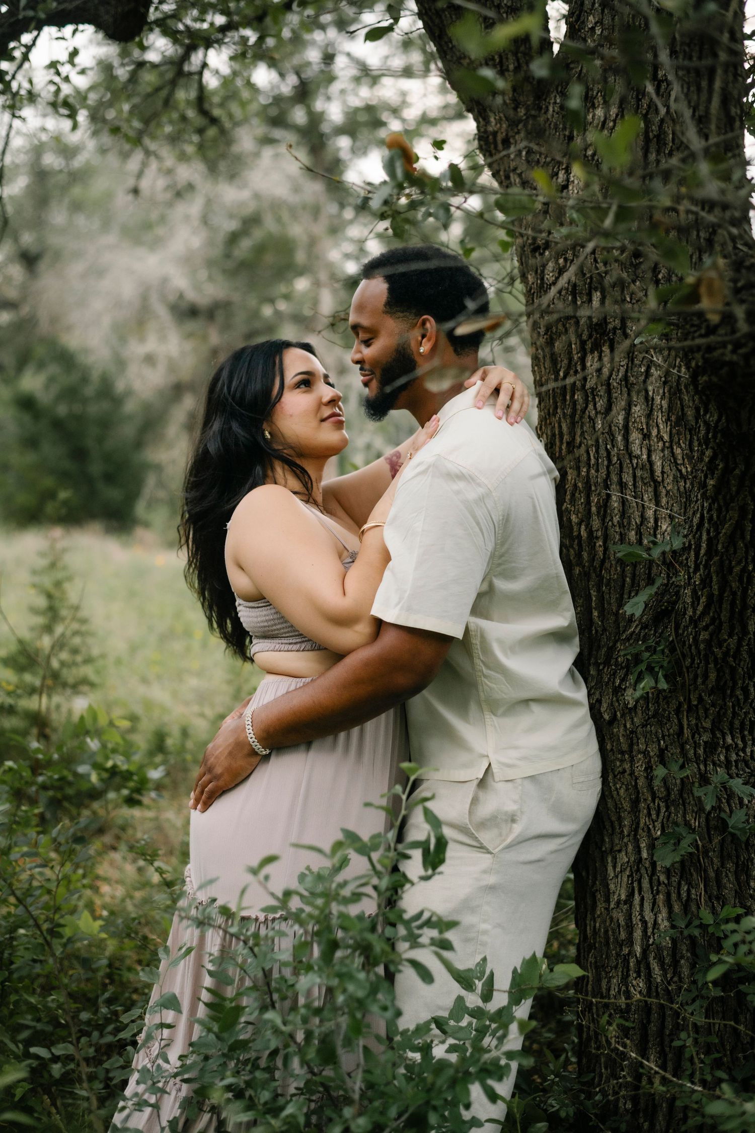 Couple embracing romantically near a tree in a lush green forest, woman in pink dress smiling up at man in cream outfit.