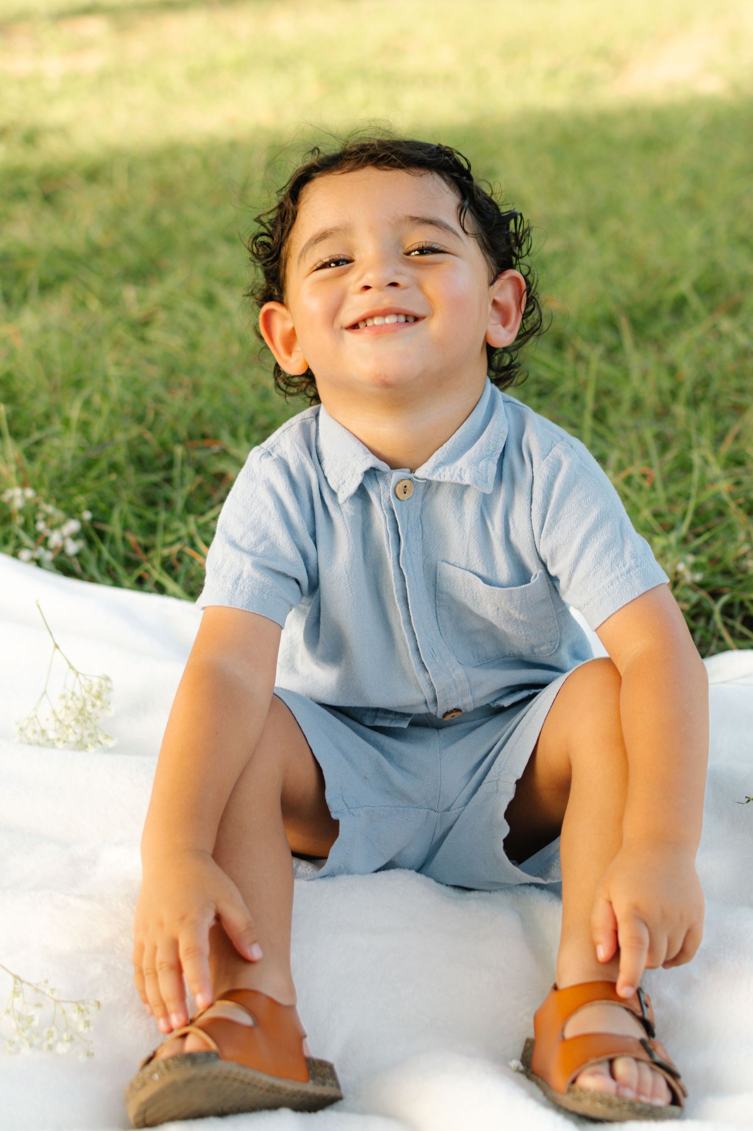 A smiling toddler boy in a light blue romper and sandals sits on a white blanket in a sunny green park.
