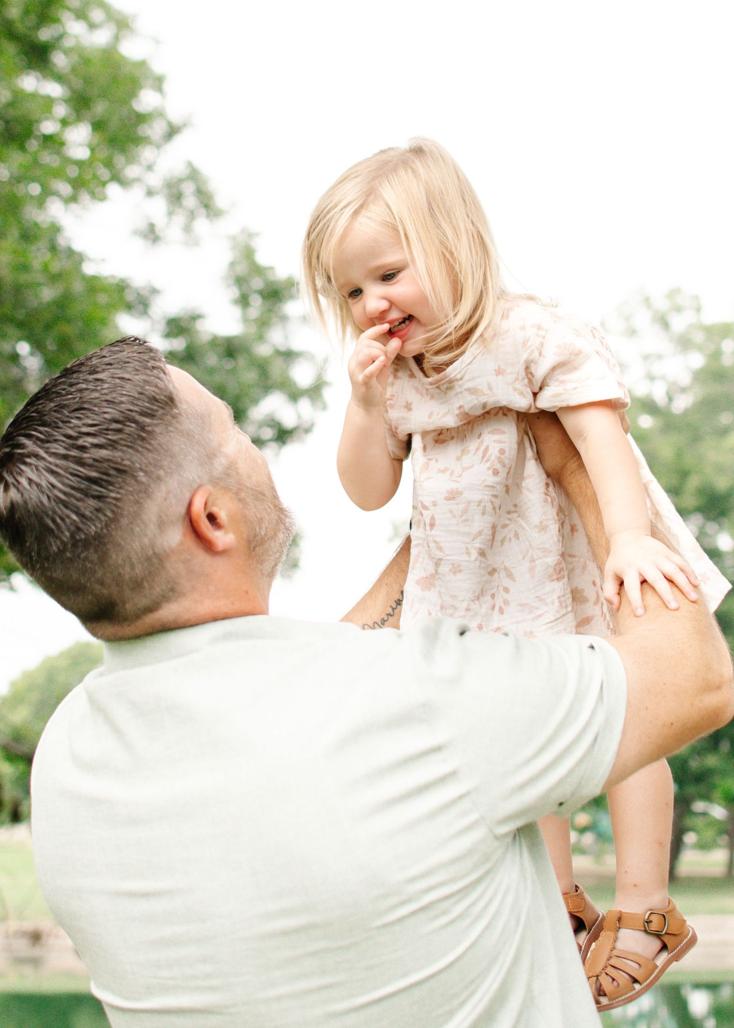 A father lifts his smiling toddler girl in a pink dress outdoors, sharing a joyful moment in a lush green park.