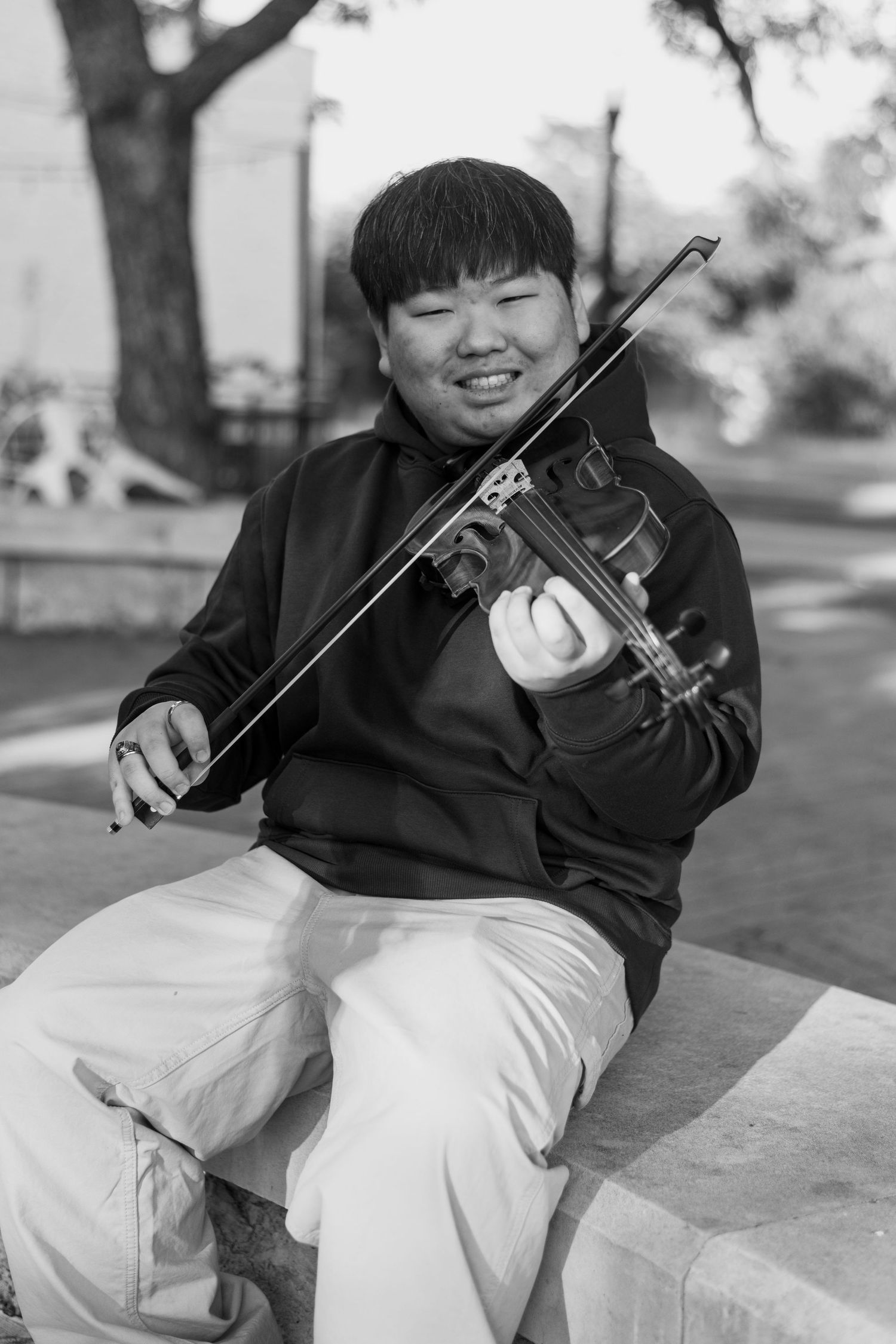Smiling young boy playing violin outdoors in black and white photo, seated on stone ledge wearing hoodie.