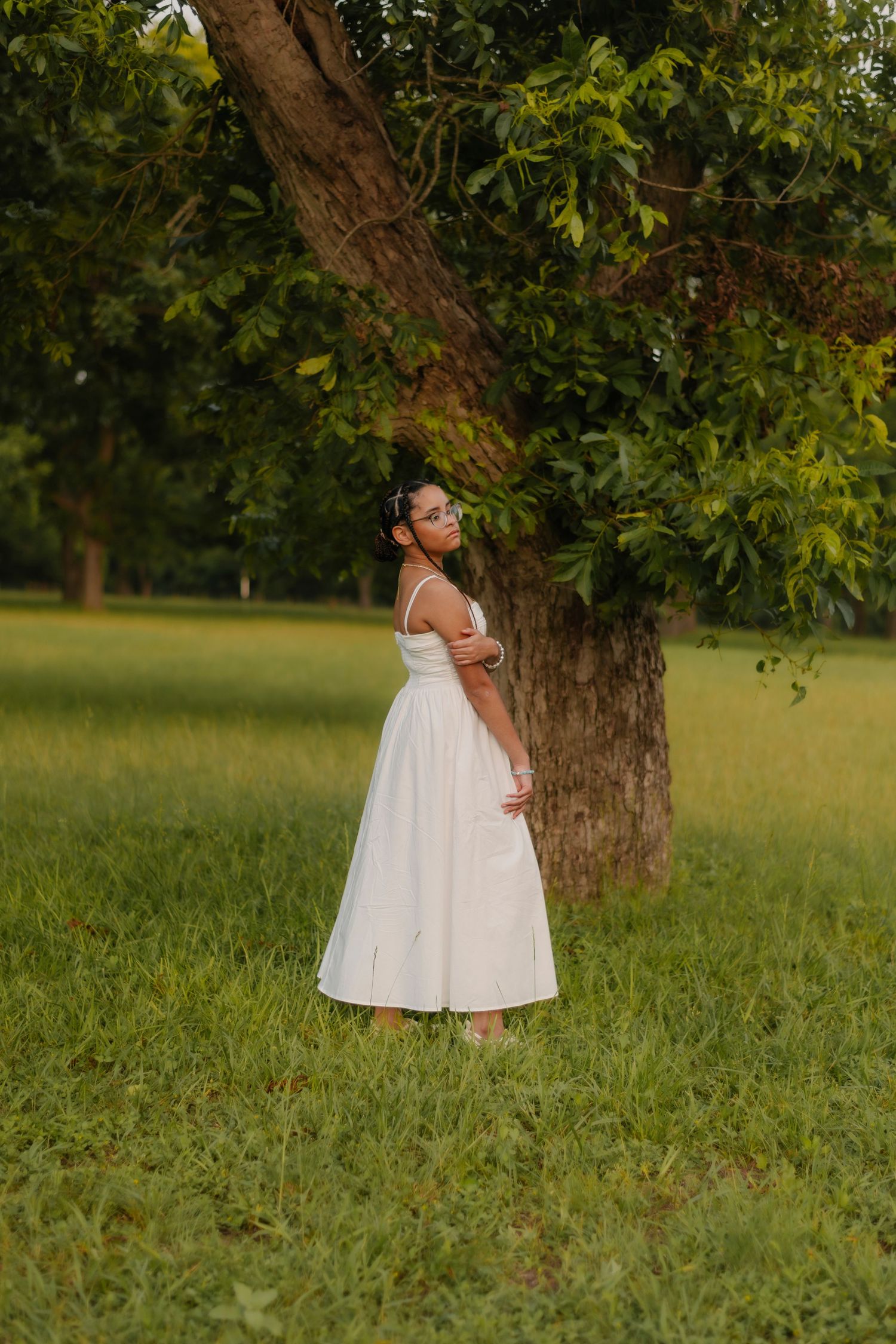 Woman in white dress standing by a large tree in a green park, looking over her shoulder in golden hour light.