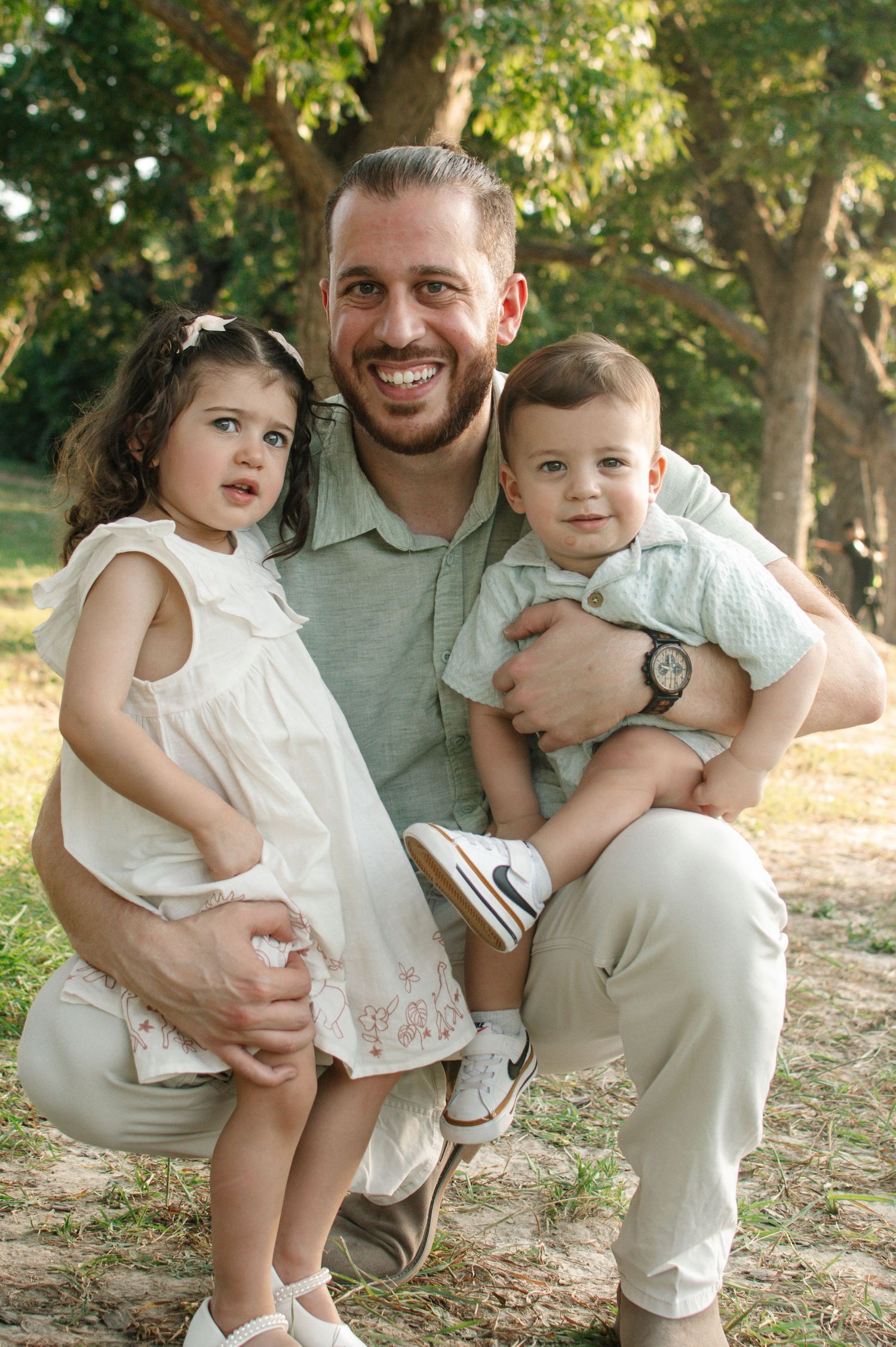 A smiling father crouches outdoors holding his baby boy and young daughter in a sunny green park.