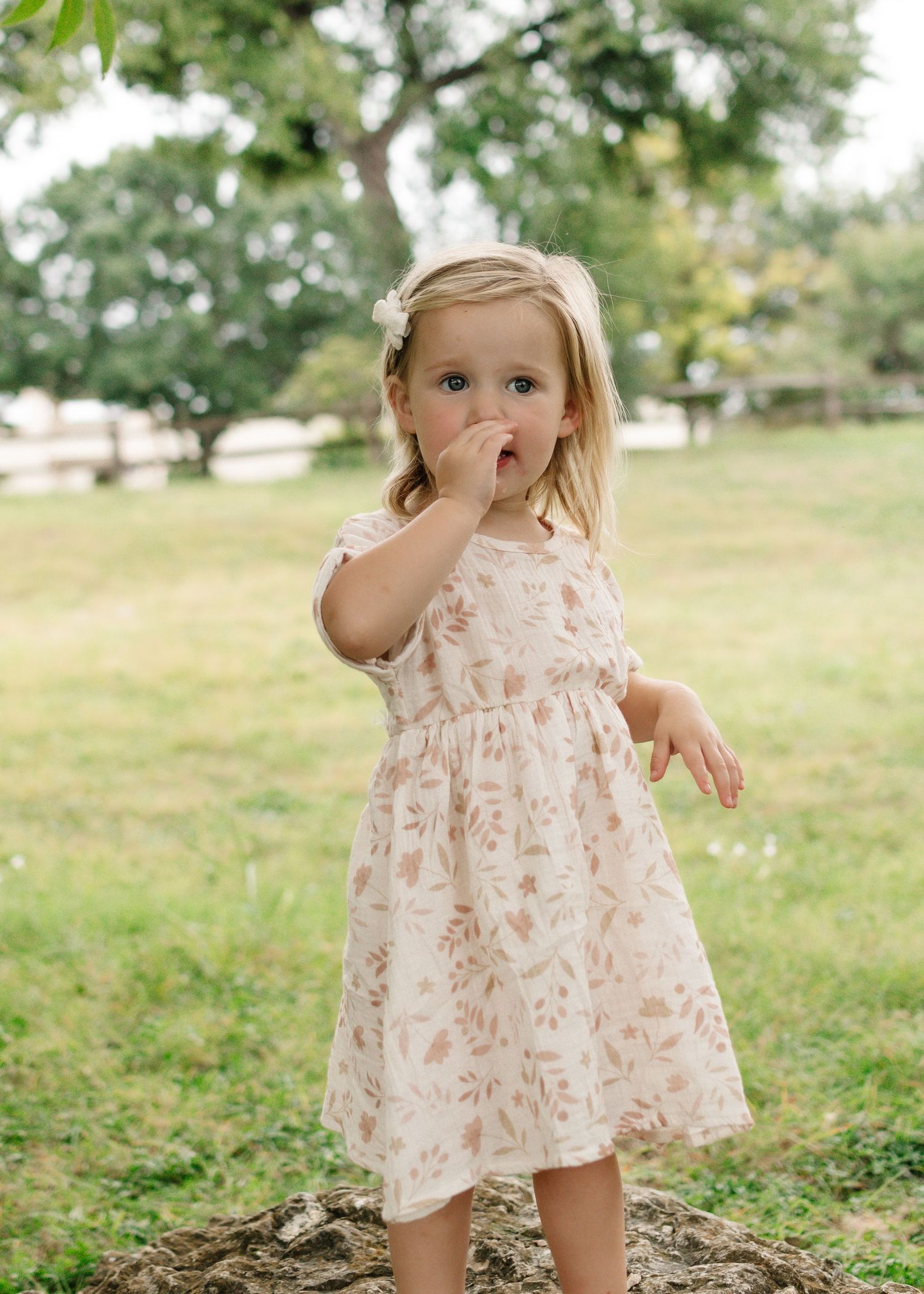 Toddler girl in a floral dress standing on a tree stump in a green park, hand near her mouth.