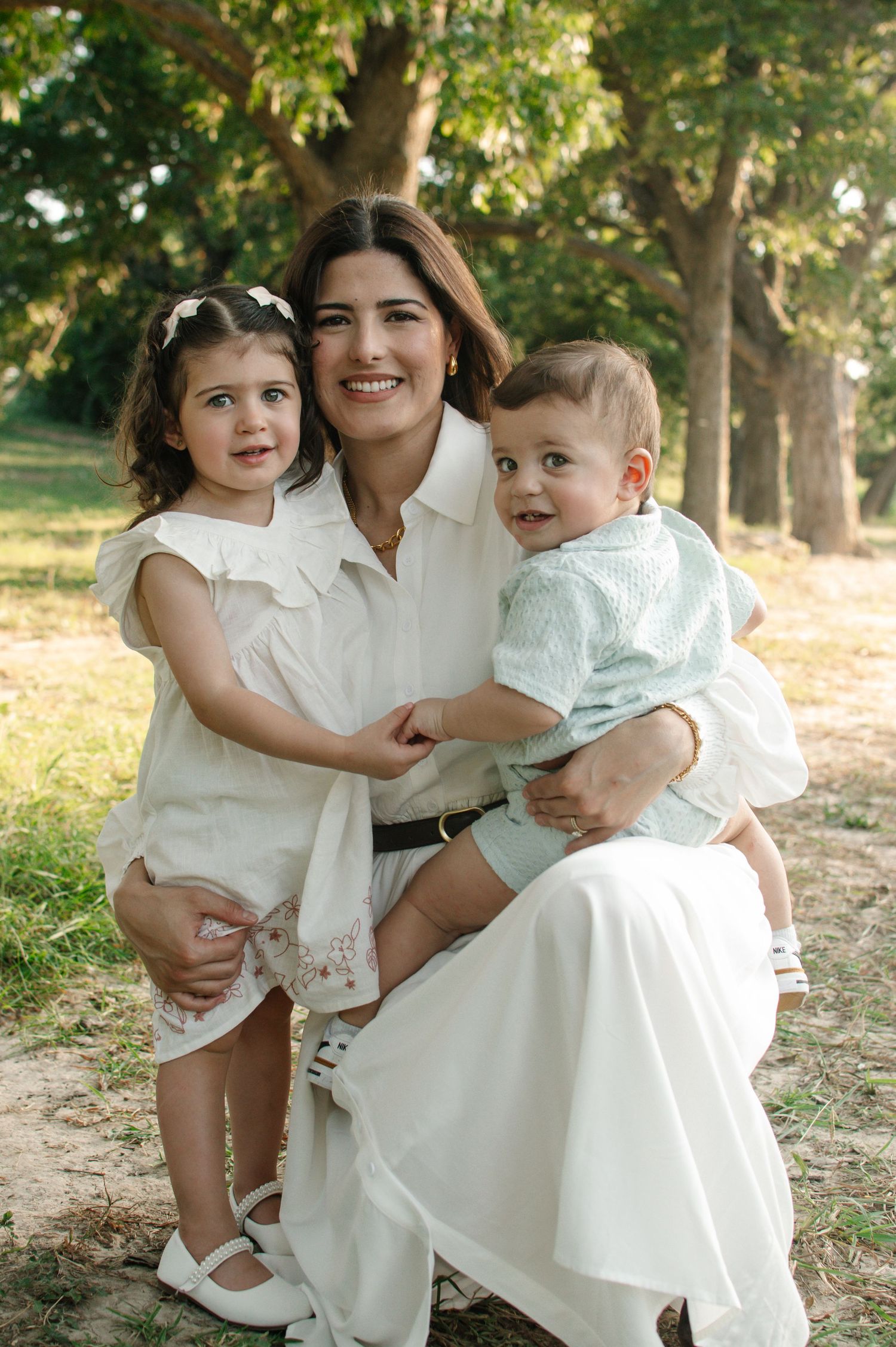 A smiling woman in white sits outdoors holding two young children, a toddler girl and baby boy, surrounded by trees.