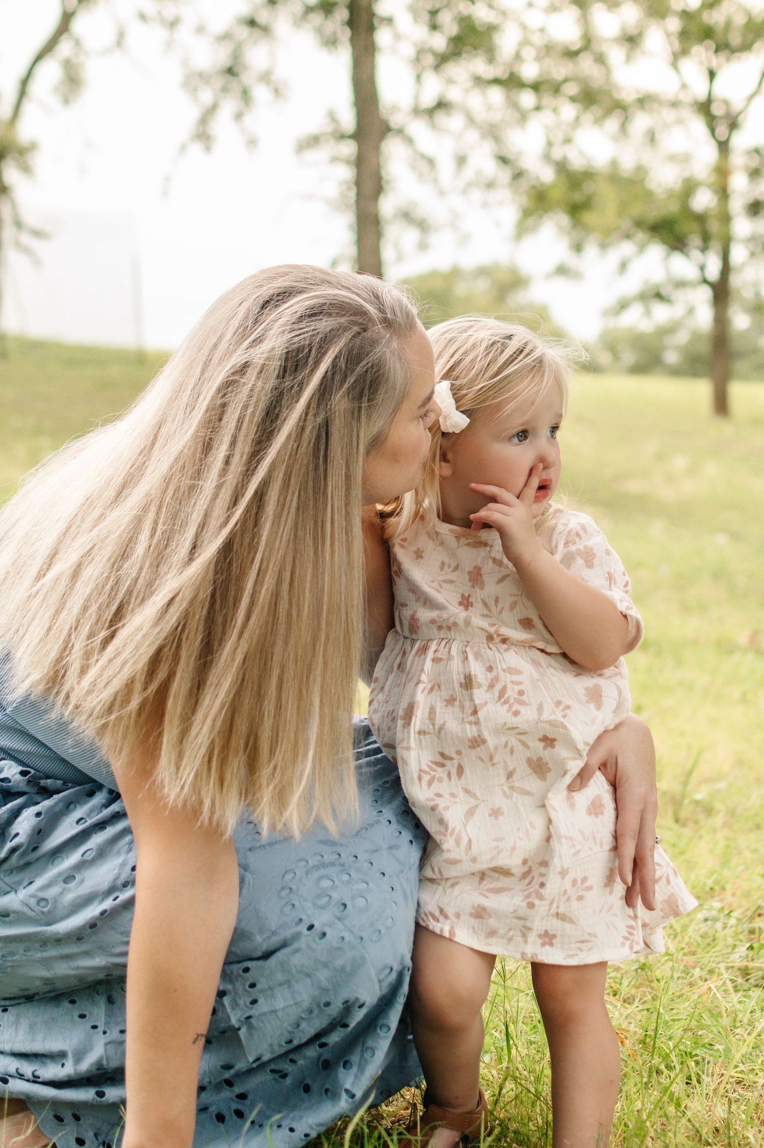 A blonde mother kisses her toddler daughter in a floral dress outdoors in a green park setting.
