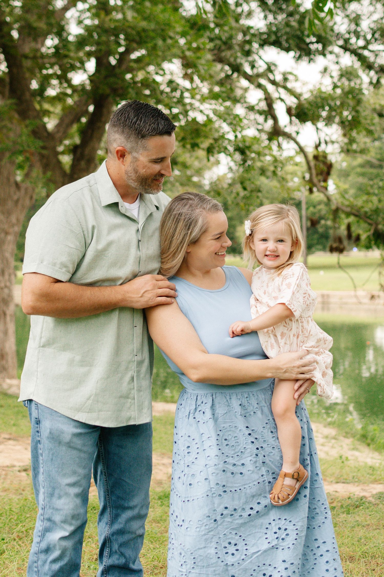 A happy family of three poses outdoors near a pond, with parents smiling at their young blonde daughter.