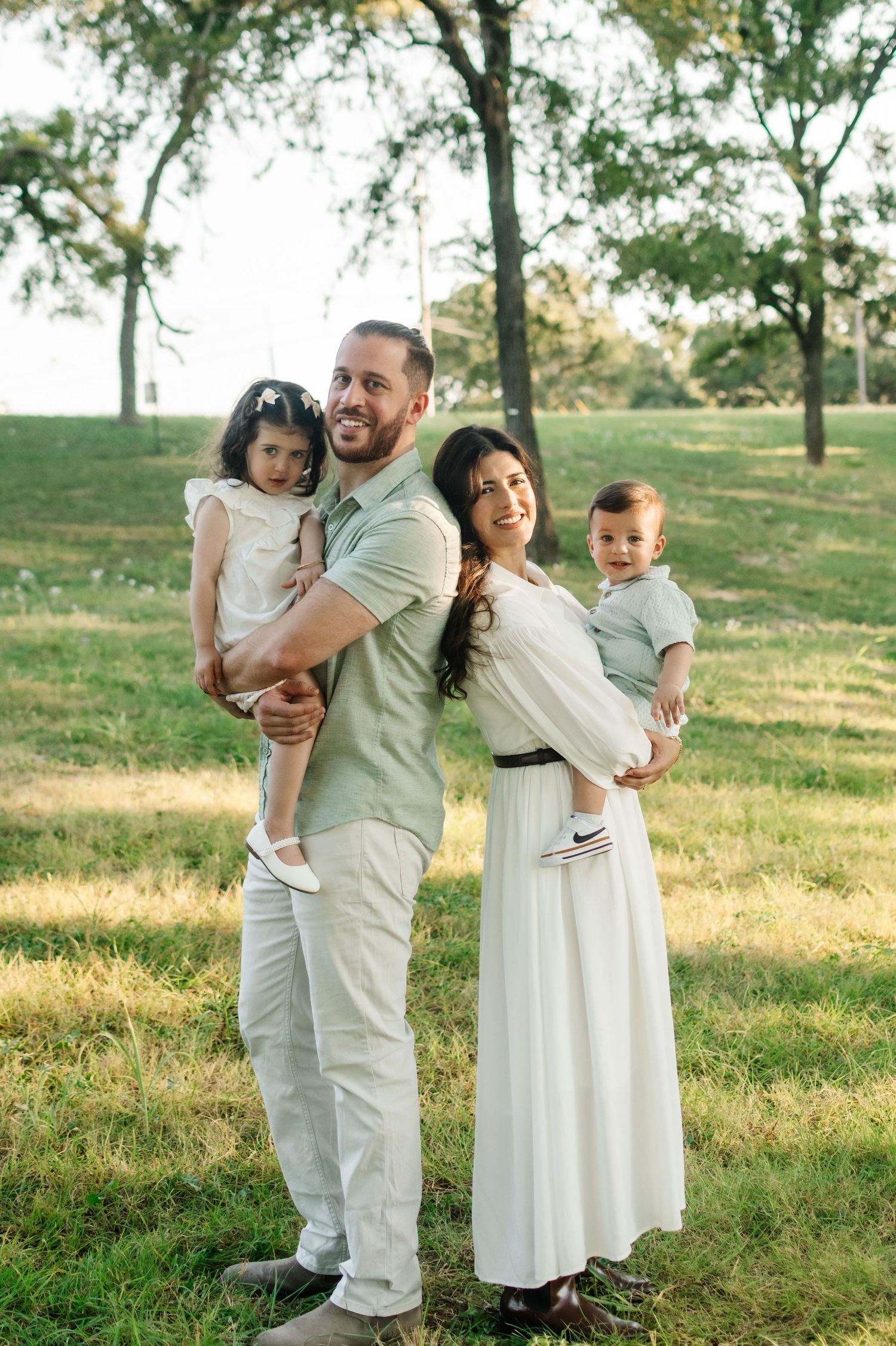 A happy family of four poses in a lush green park, parents holding two young children in coordinated neutral outfits.