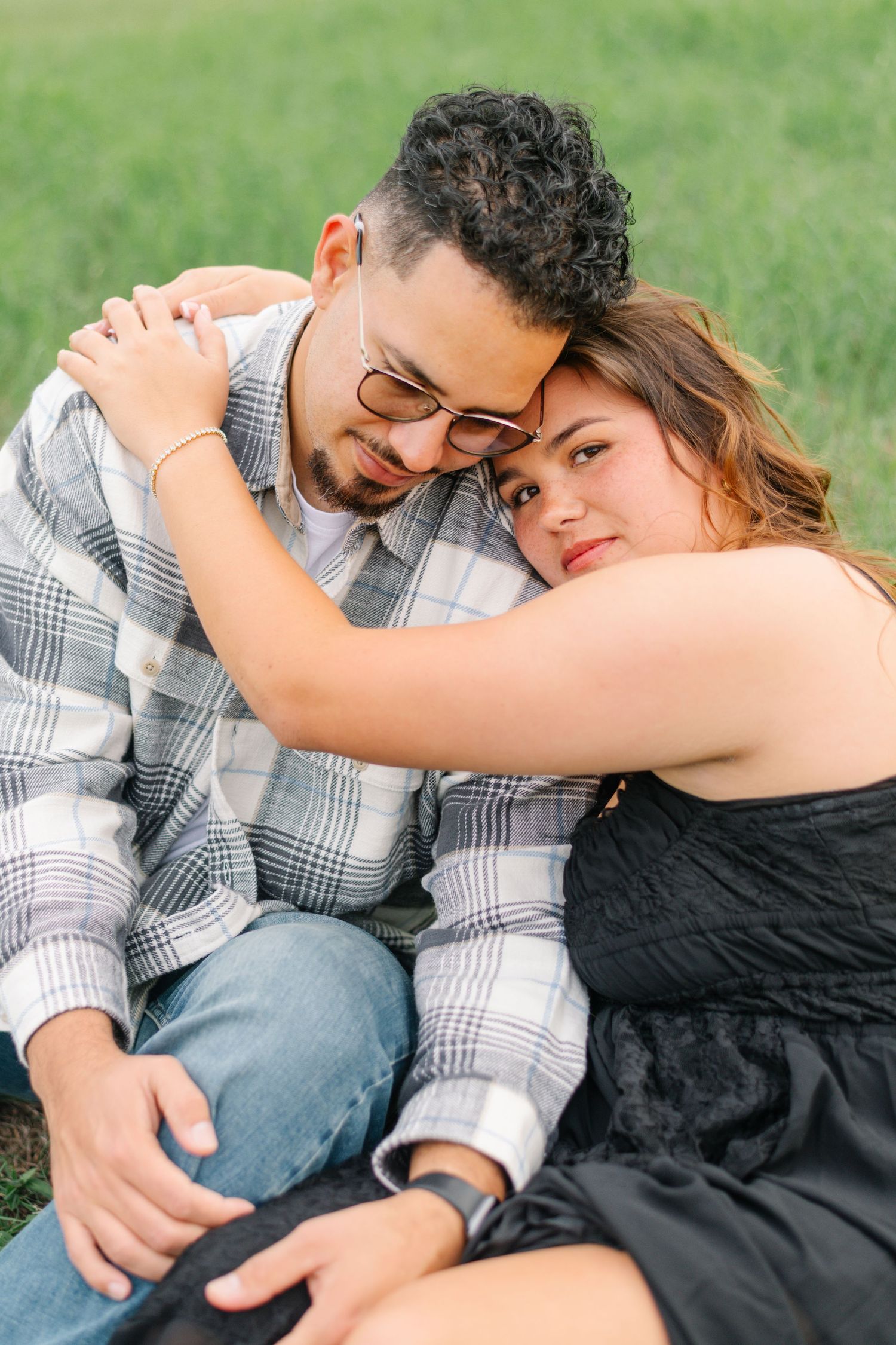 A couple sits together on grass, the woman in a black dress leaning on a man in a plaid shirt and sunglasses.
