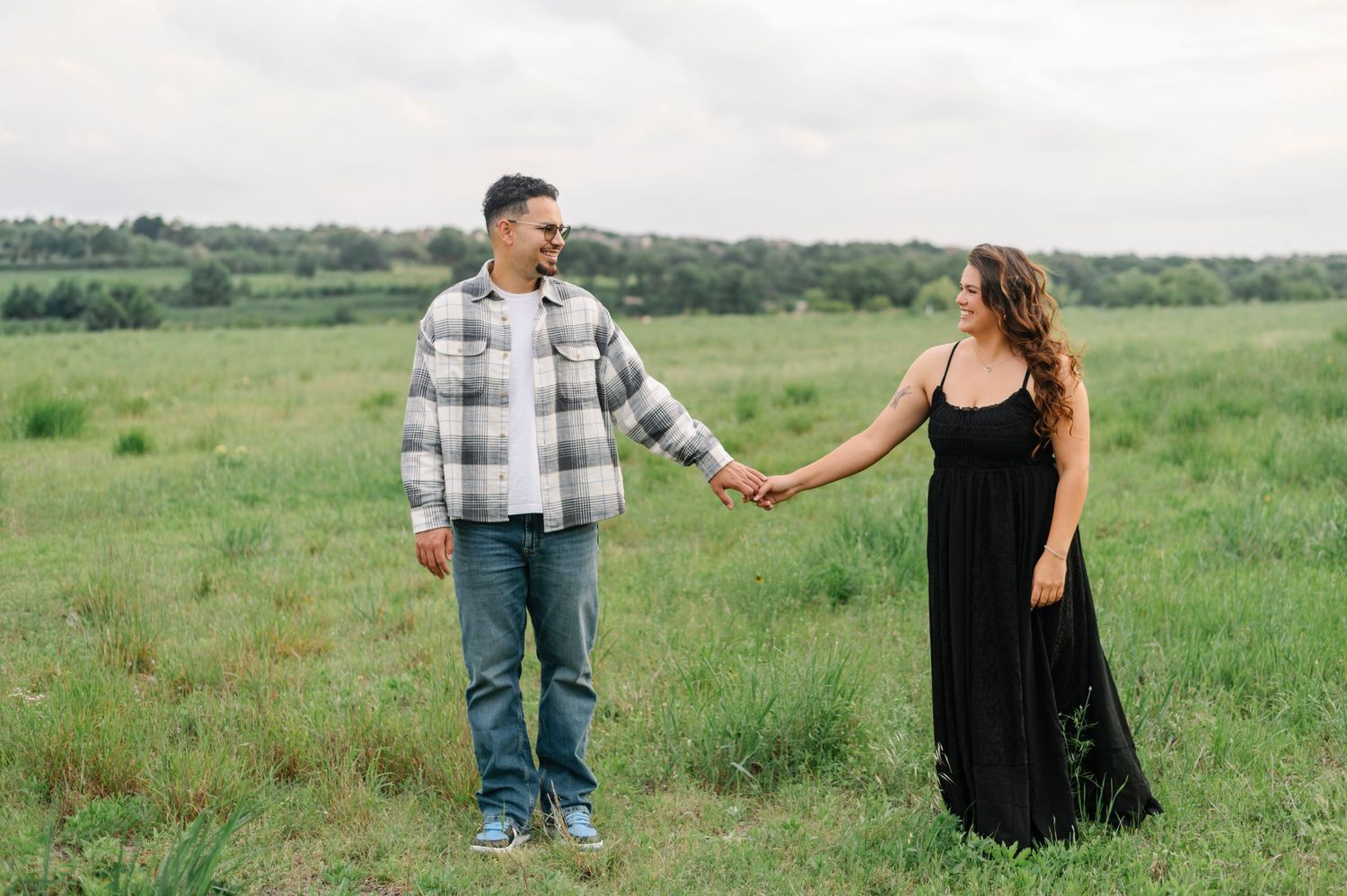 Couple holding hands in a green field, man in plaid shirt and jeans, woman in black maxi dress, overcast sky.