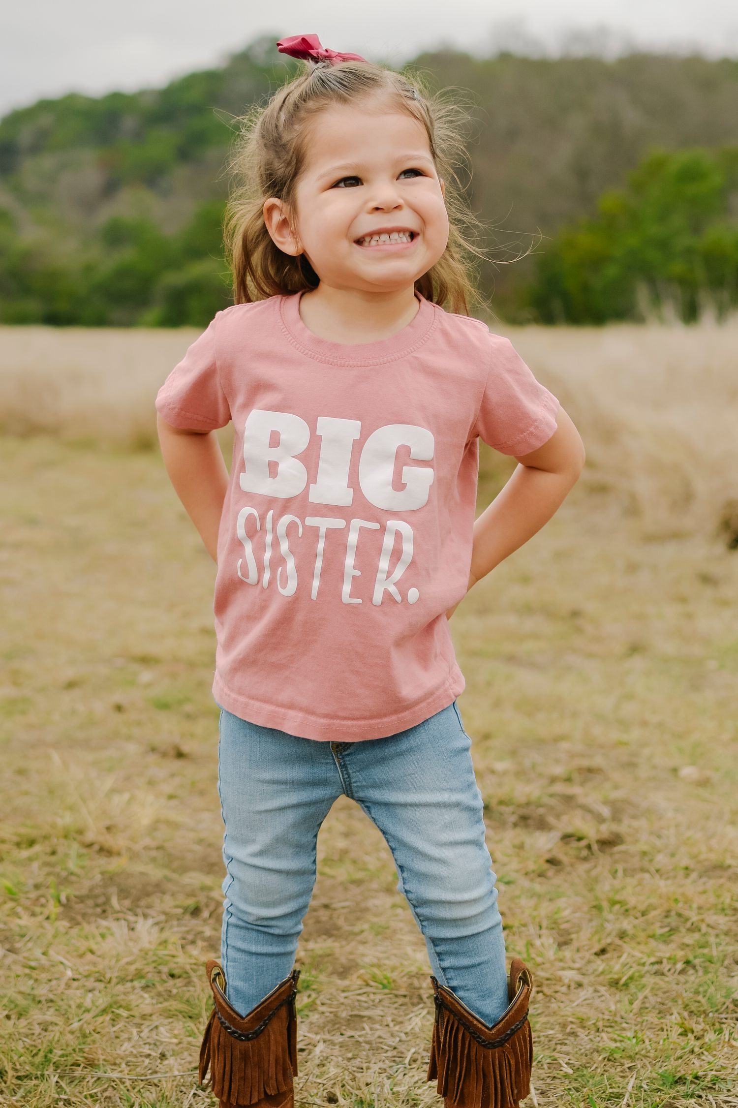 Smiling toddler girl wearing a pink Big Sister t-shirt and jeans with fringe boots, posing outdoors in a field.