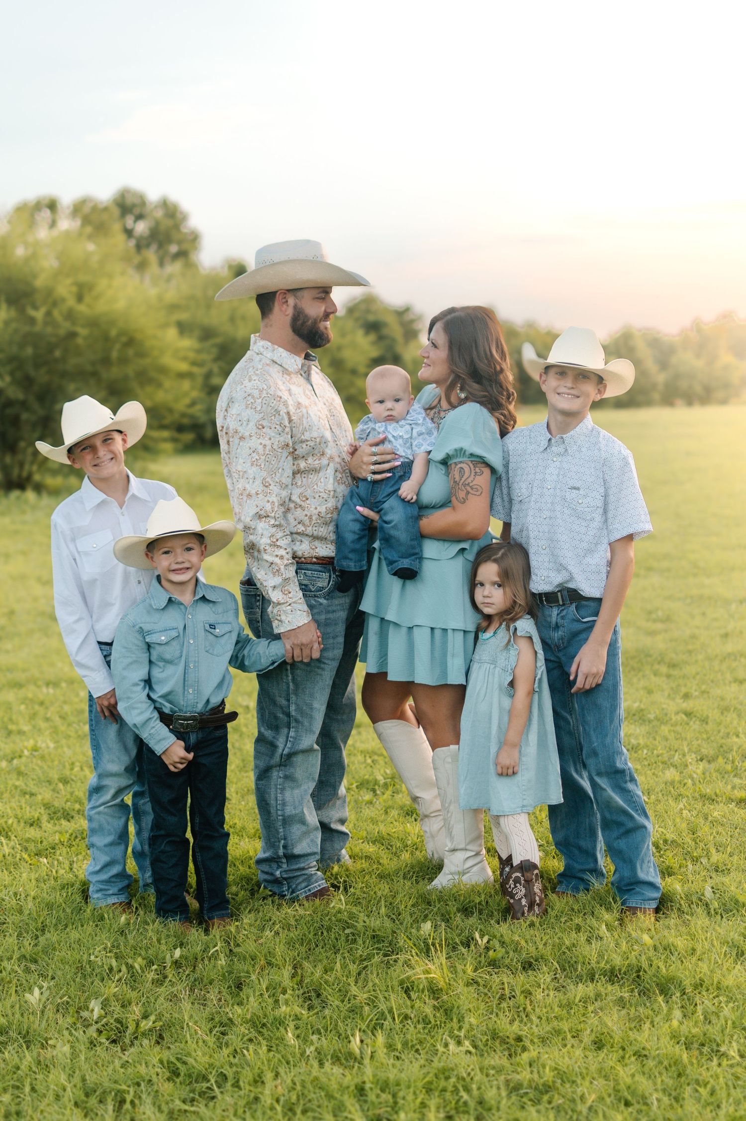 Family of seven in cowboy hats posing in a green field at golden hour, wearing coordinated western outfits.