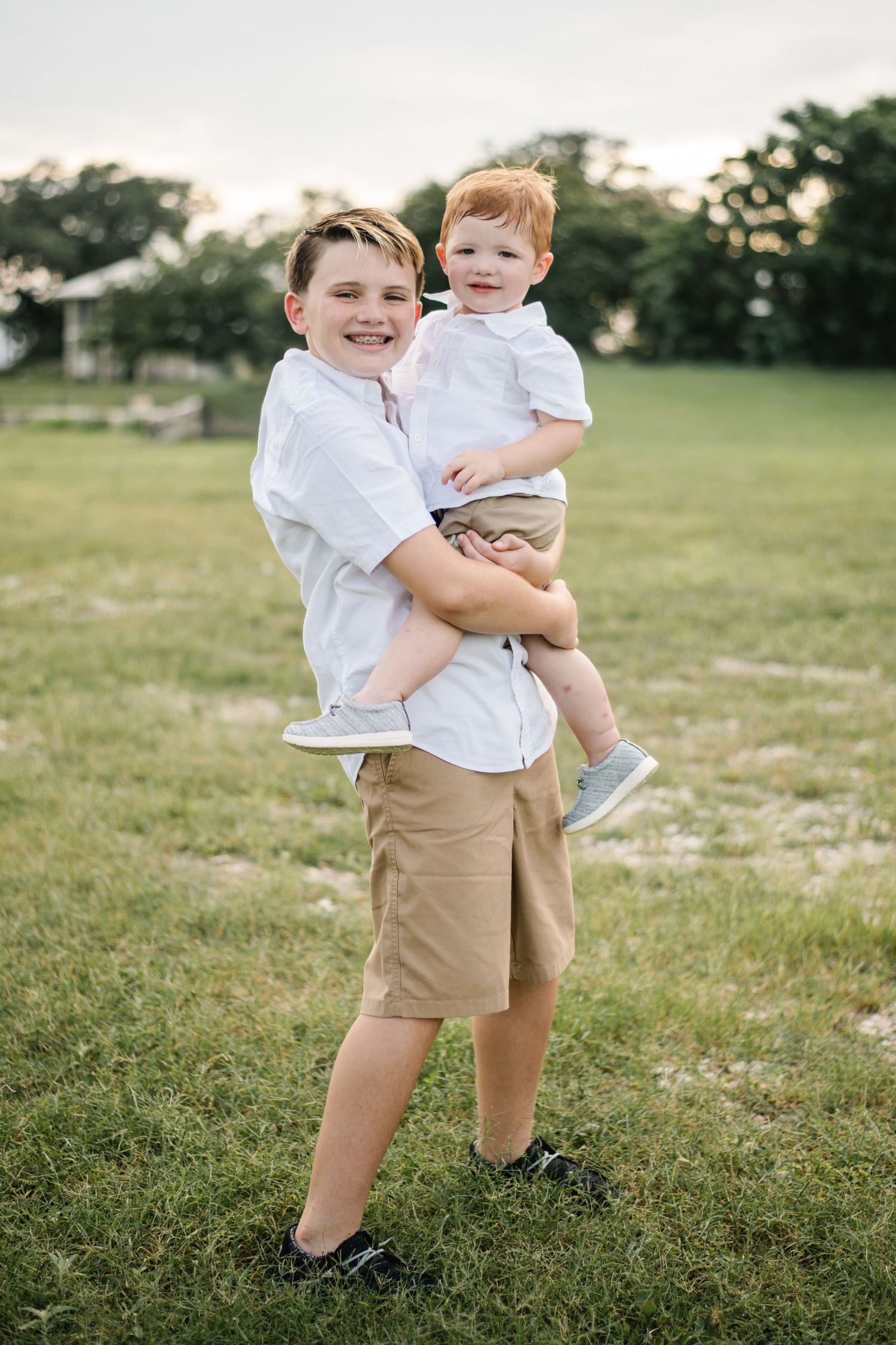 Two boys in matching white shirts and khaki shorts pose outdoors, older brother holding younger toddler sibling in a grassy field.