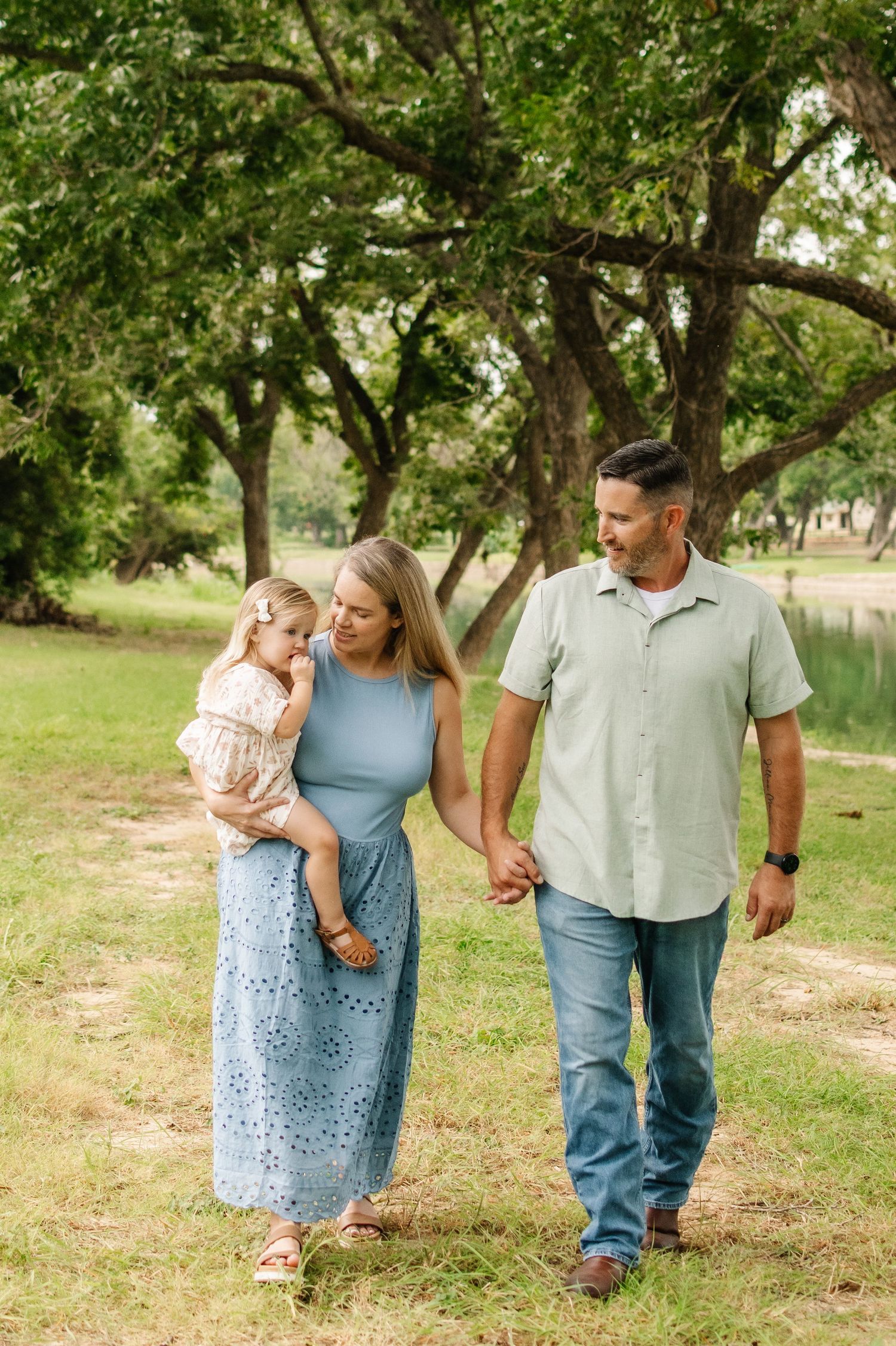 A family of three walks through a lush green park, mother holding toddler while father holds her hand.