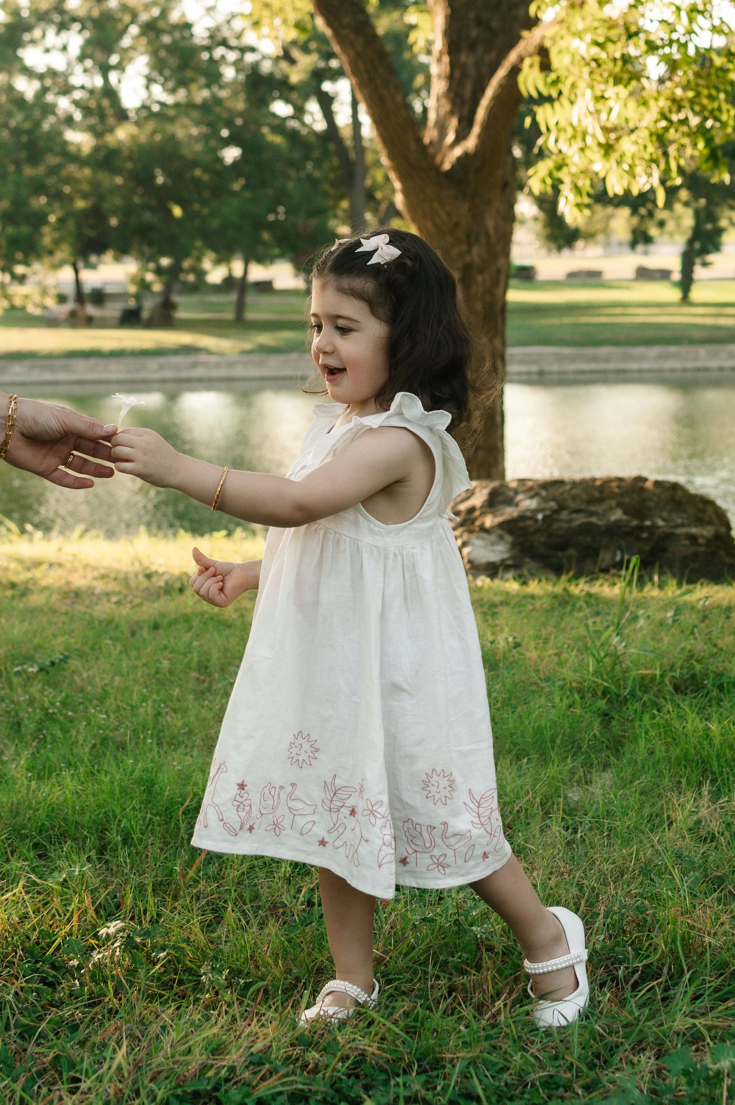 A joyful toddler girl in a white embroidered dress reaches out while playing in a lush green park.