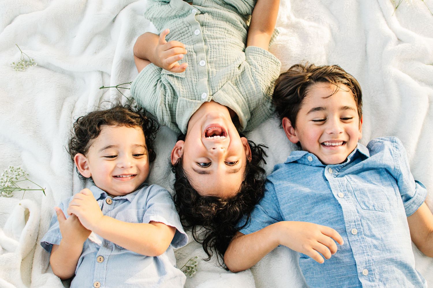 Three happy children lying on a white blanket outdoors, laughing and smiling together on a sunny day.