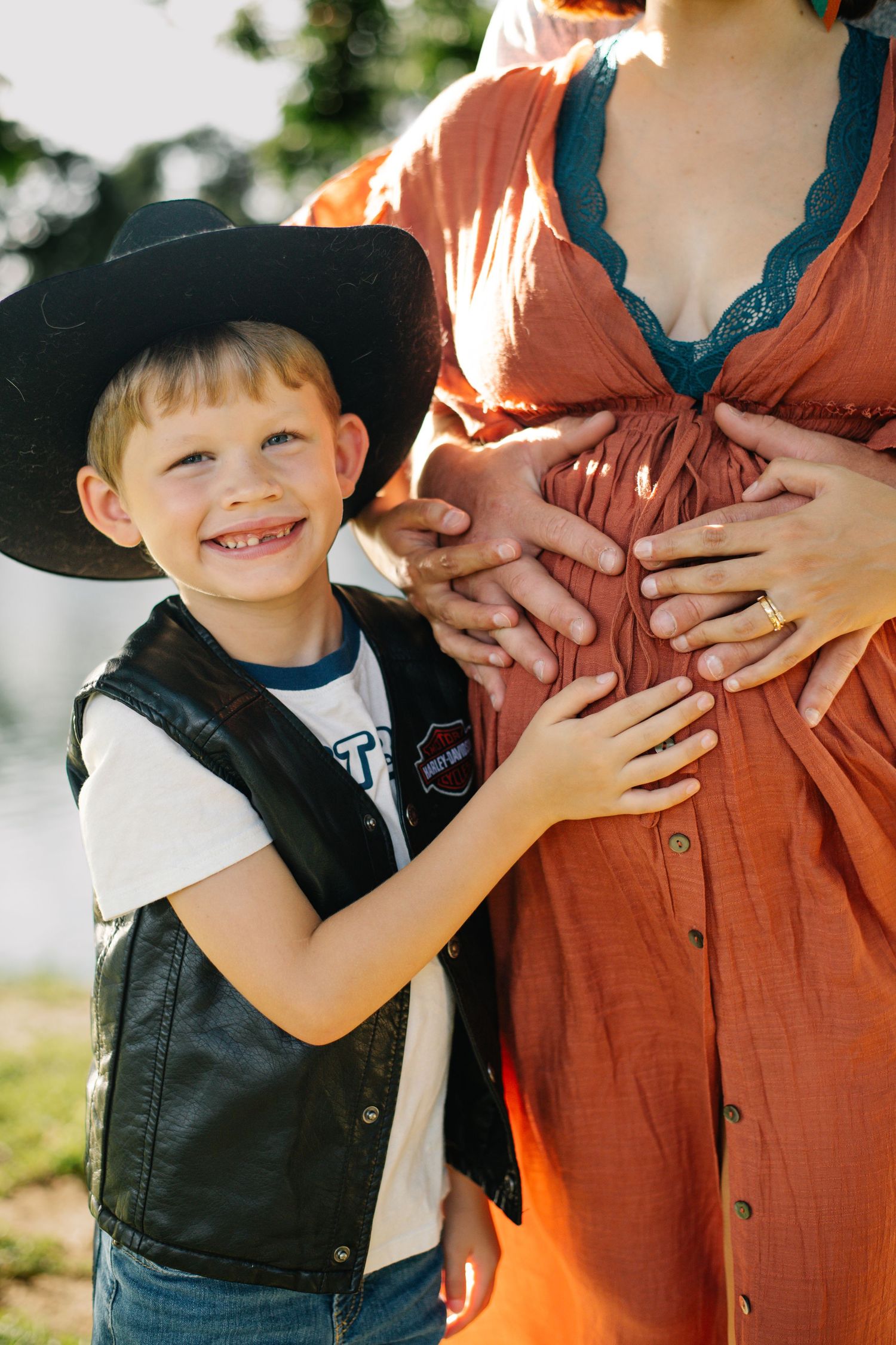Smiling boy in cowboy hat and leather vest touching pregnant woman's belly in outdoor maternity photo.