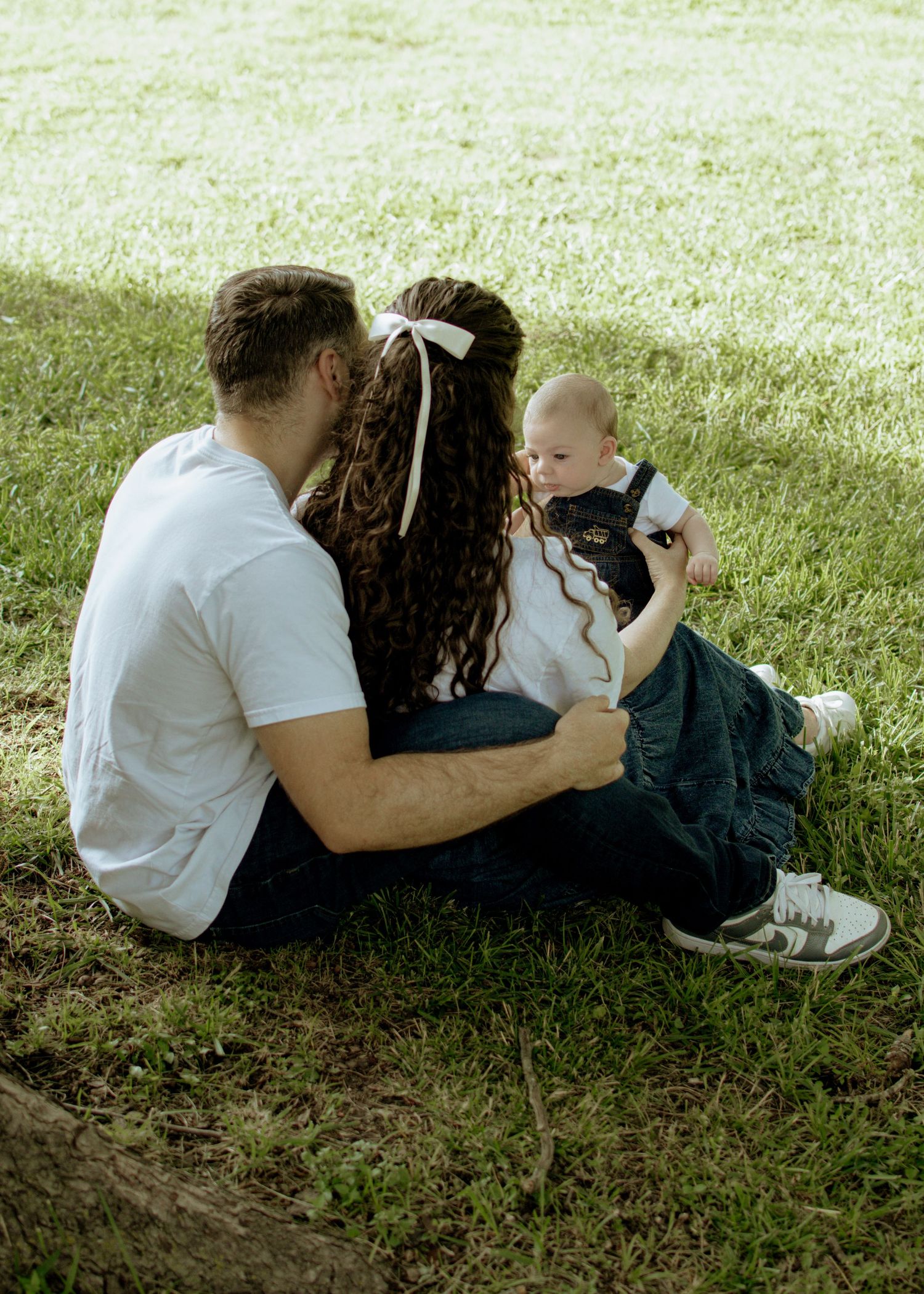 Family sitting together on grass while holding their baby in casual outdoor setting.