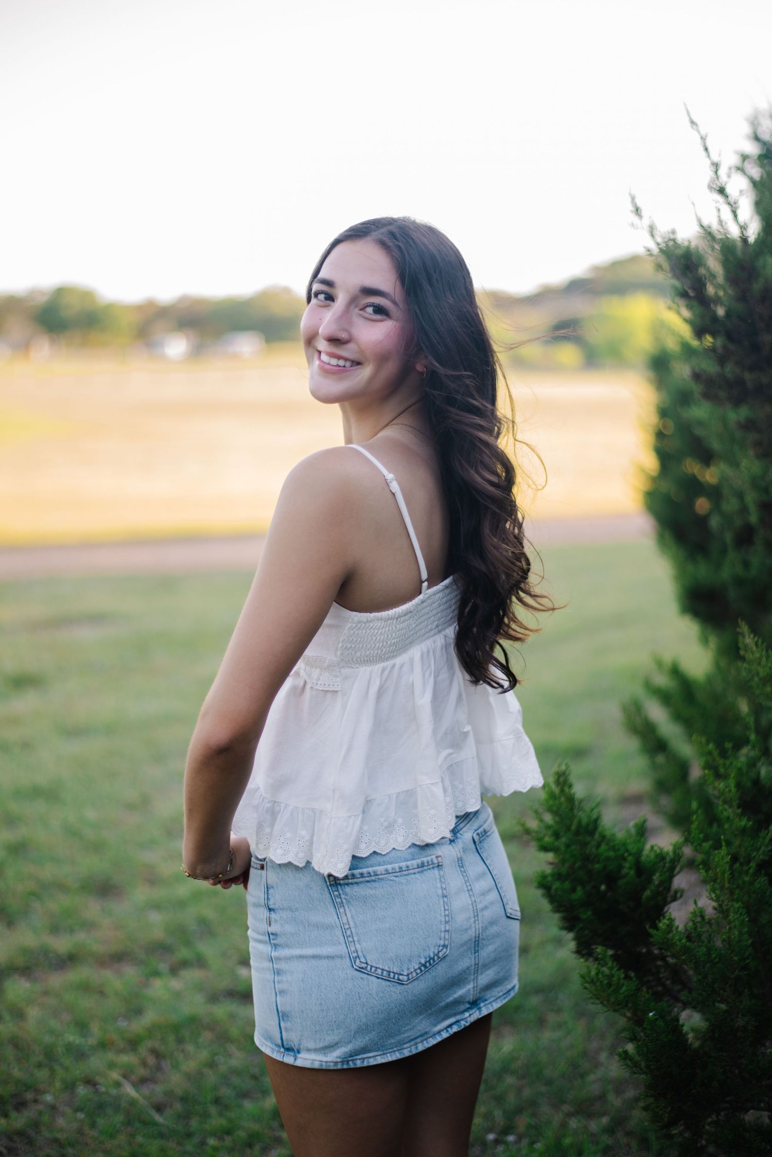 A woman in a white ruffle top and denim skirt poses outdoors in a natural setting.