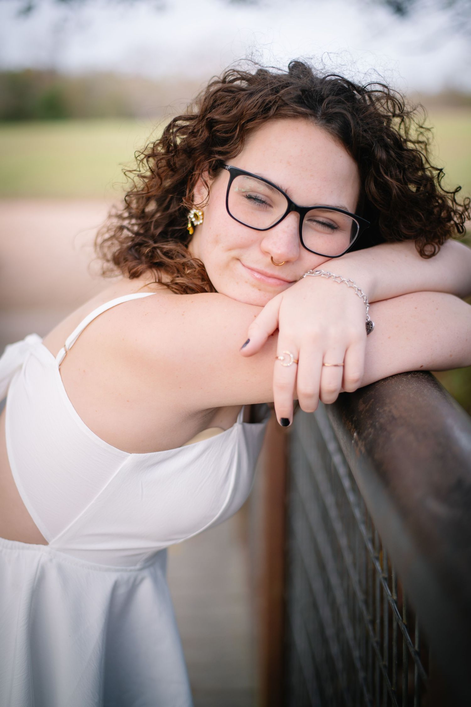 Artistic portrait series showing someone with curly hair and glasses in white top resting against a railing in soft focus.