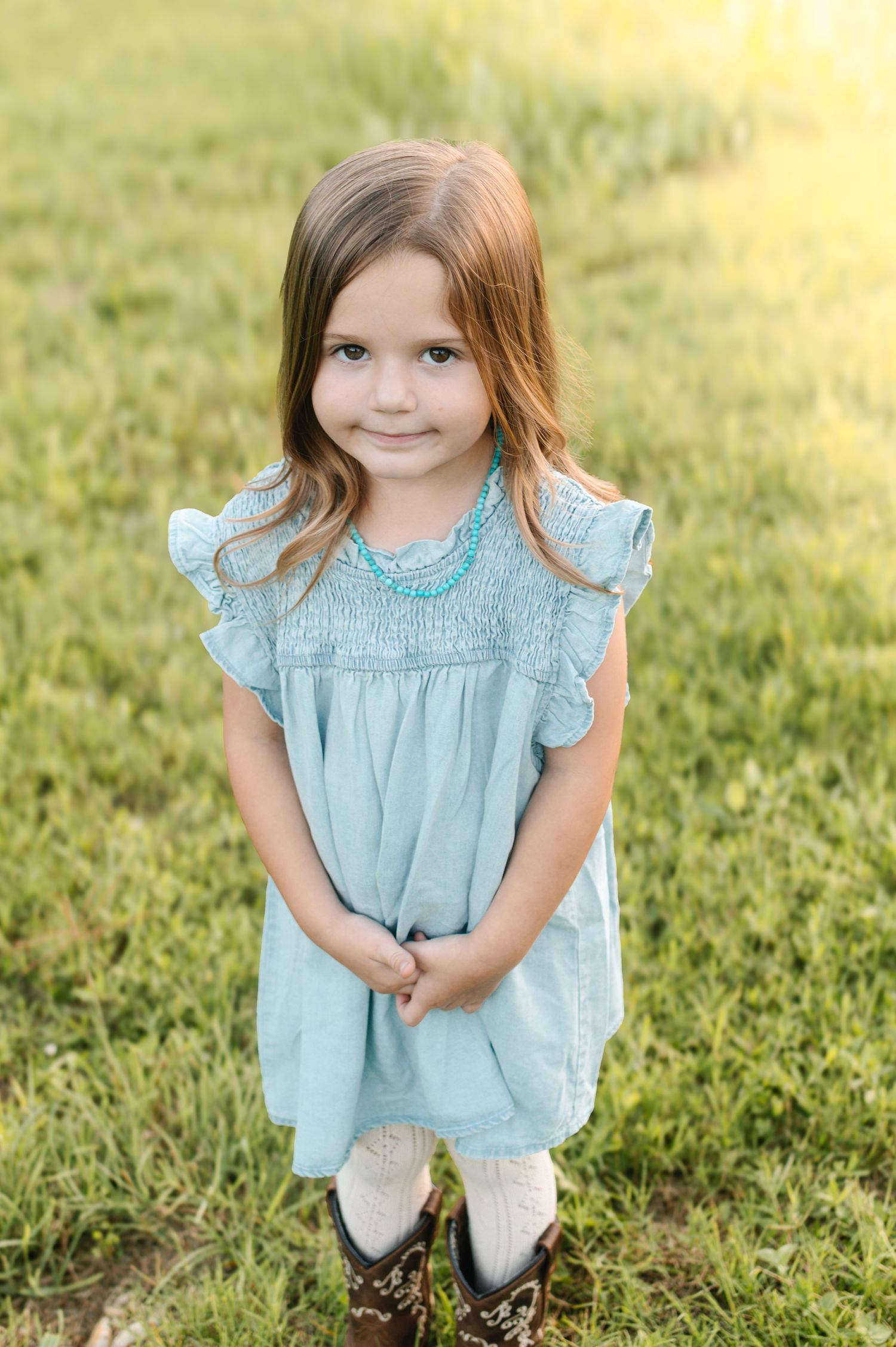 Young girl in light blue dress with turquoise necklace and cowboy boots standing on green grass.