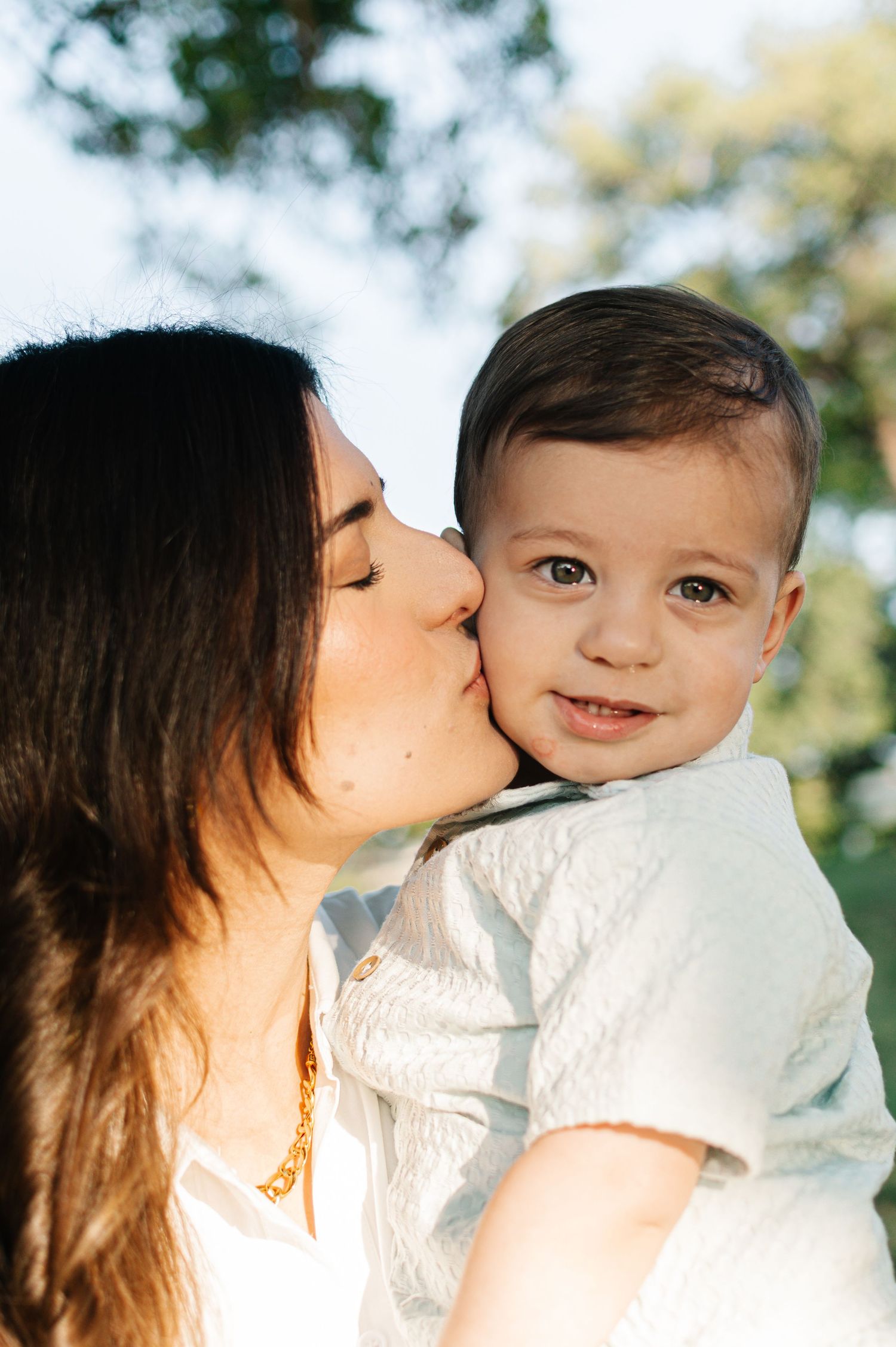 A loving mother kissing her smiling baby boy on the cheek outdoors in soft natural light.