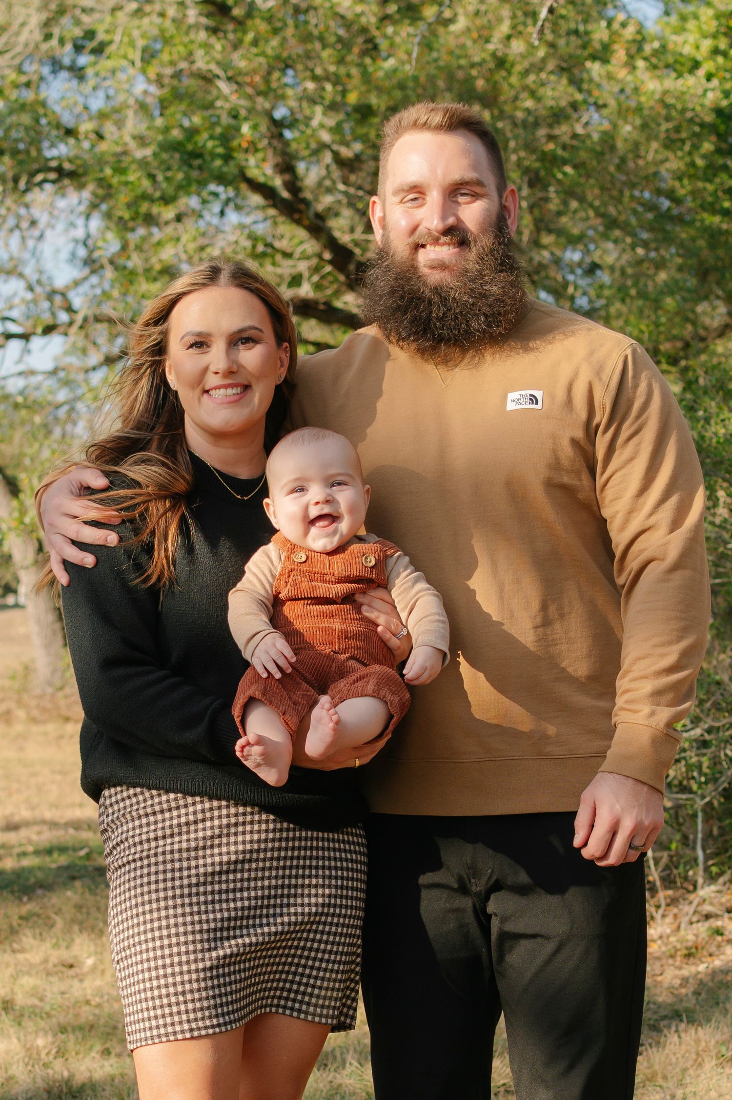 Happy family portrait outdoors with smiling couple holding adorable baby in rust overalls among autumn trees.