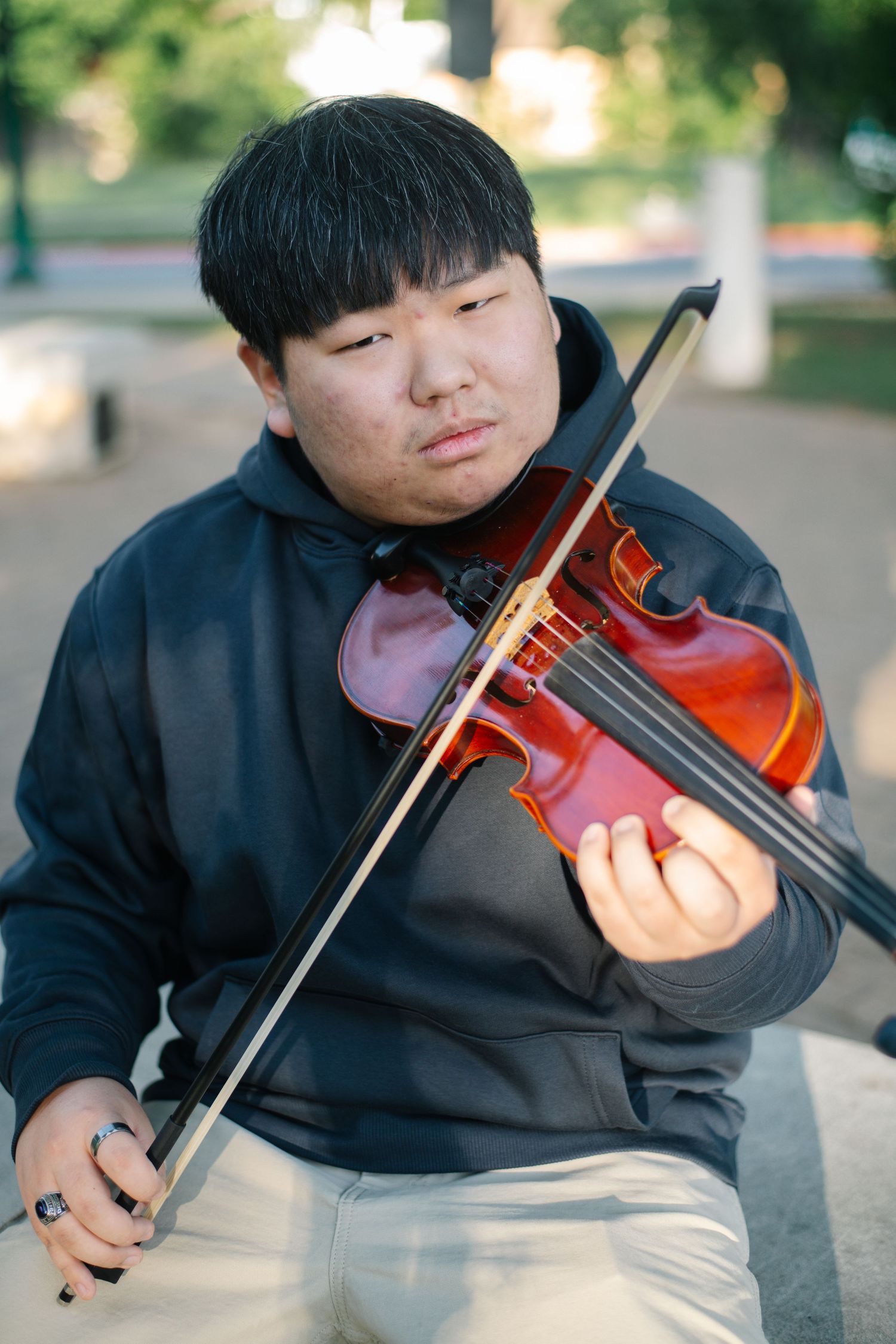 Young Asian man playing violin outdoors, wearing a dark hoodie, focused on his performance in a park setting.