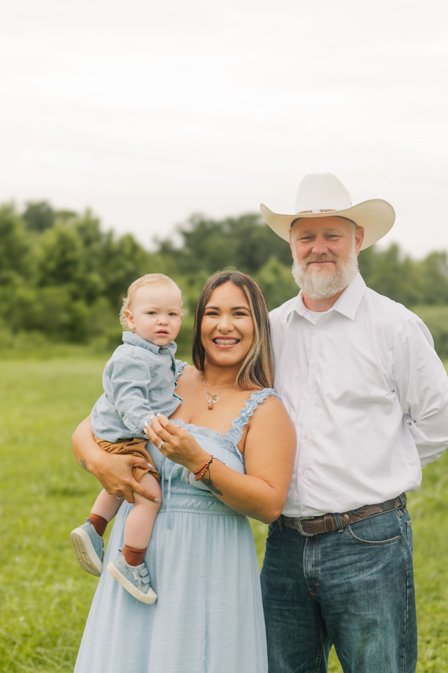 A smiling family poses outdoors, with a woman in a blue dress holding a baby and a man in a cowboy hat beside her.