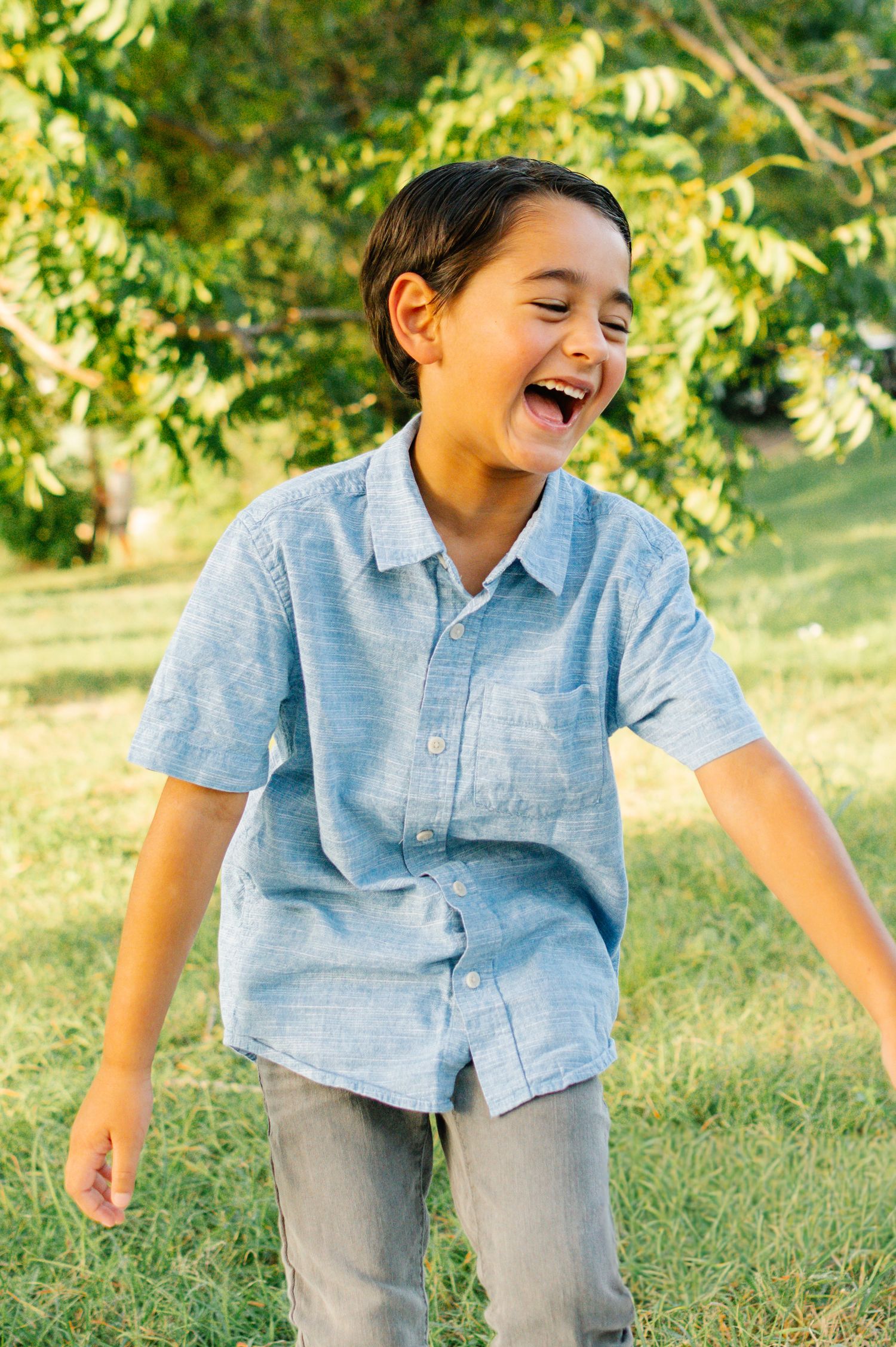 A joyful young boy in a light blue shirt laughs outdoors in a sunny green park.