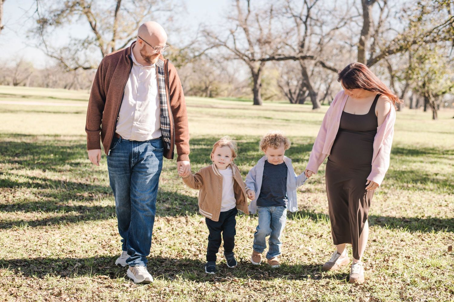 Family of four walking hand-in-hand outdoors in a sunny park, parents with two young toddlers enjoying a fall day.