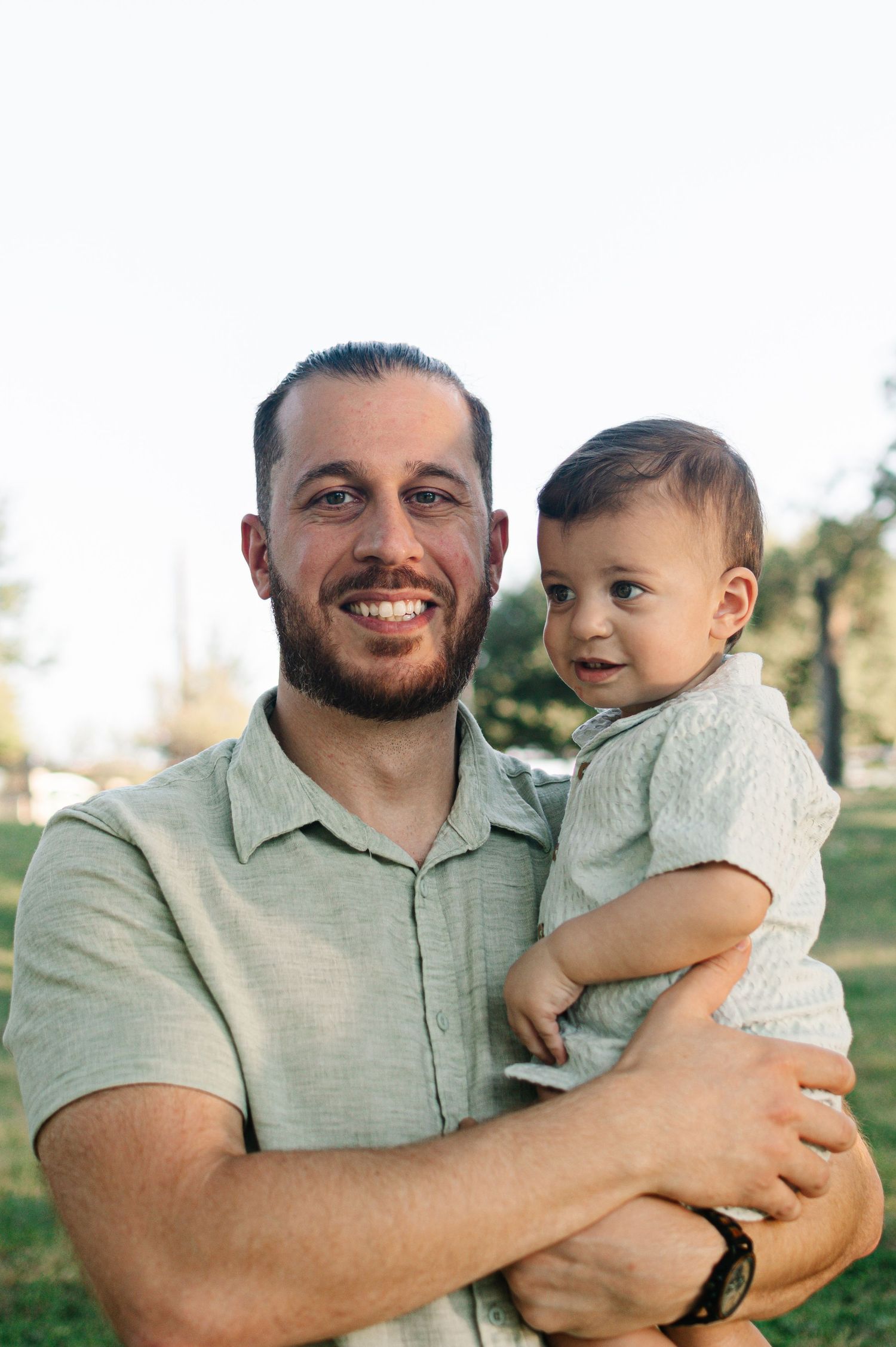 Smiling man with beard holds a happy baby boy outdoors in a sunny park setting.