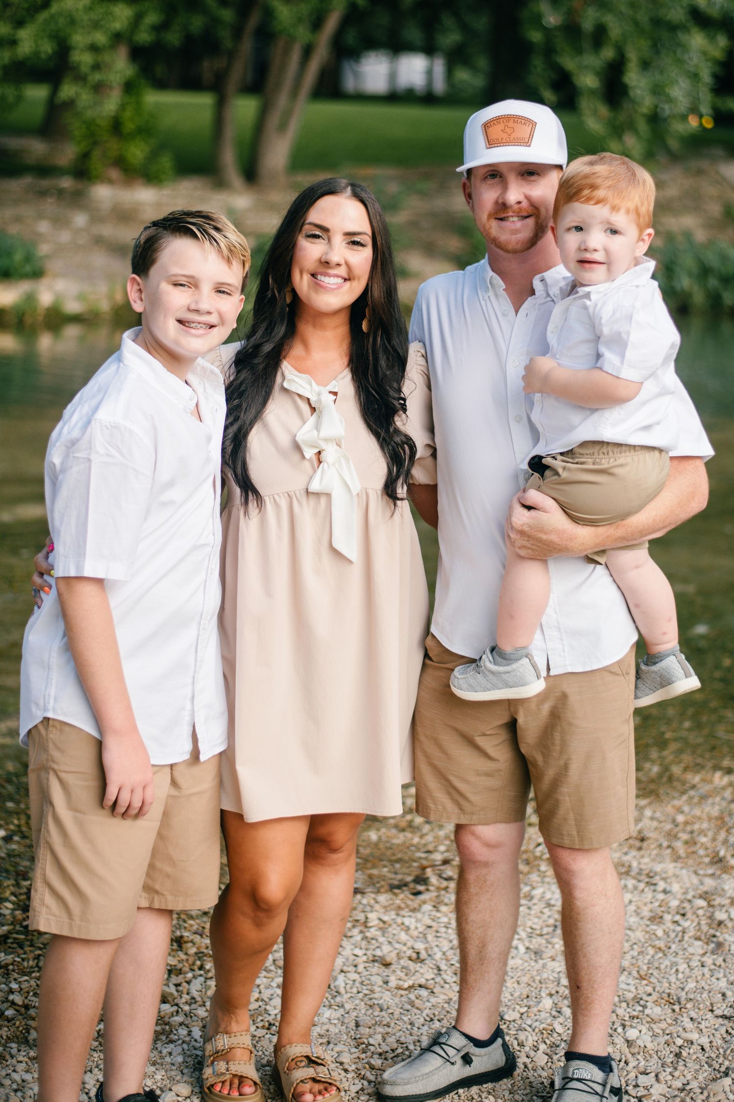 Happy family of four posing outdoors near a river, wearing coordinated white and khaki outfits on a sunny day.