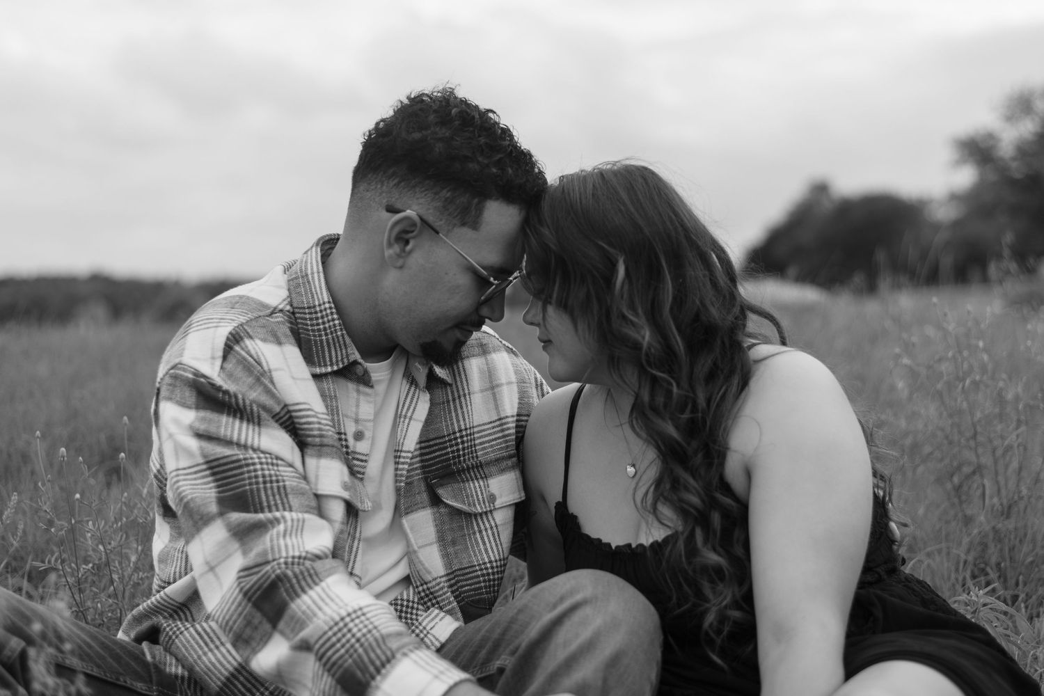 Black and white photo of a romantic couple sitting in a field, leaning foreheads together tenderly.