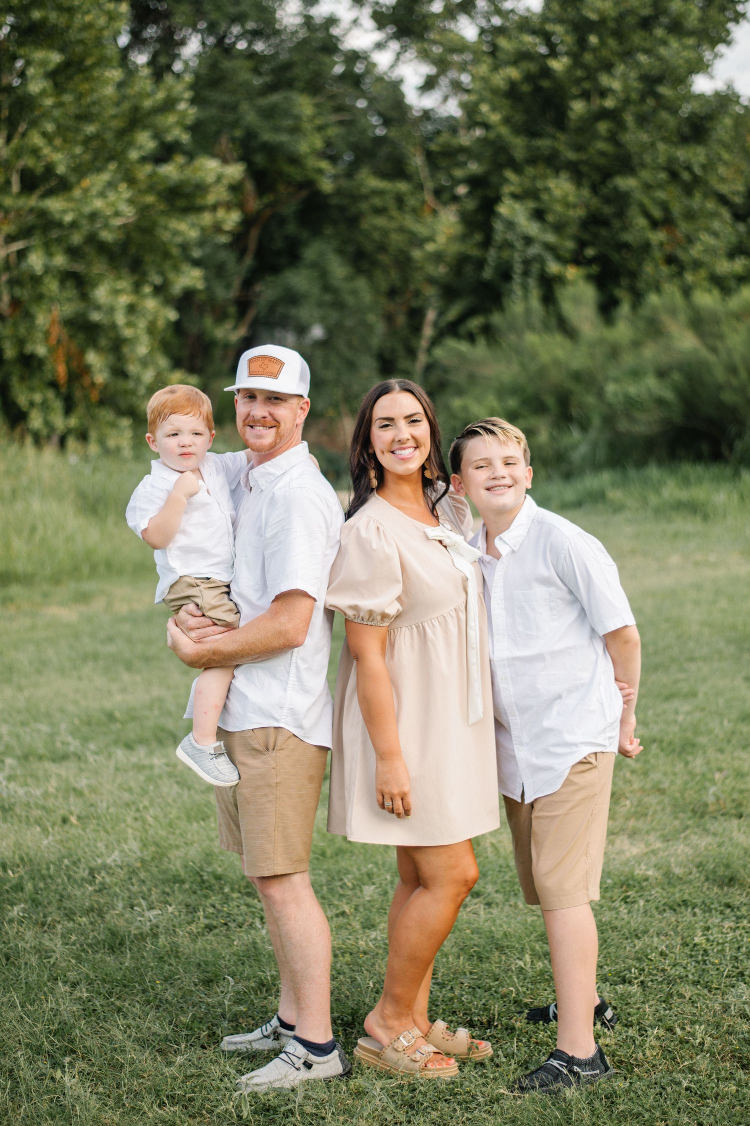 Happy family of four posing outdoors in coordinating neutral outfits on a green lawn with trees behind them.