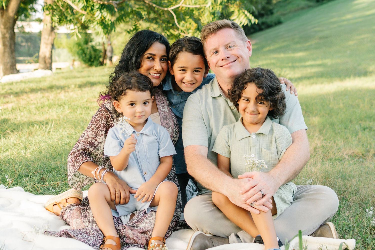 Happy family of five sitting on a blanket in a green park, smiling together on a sunny day.