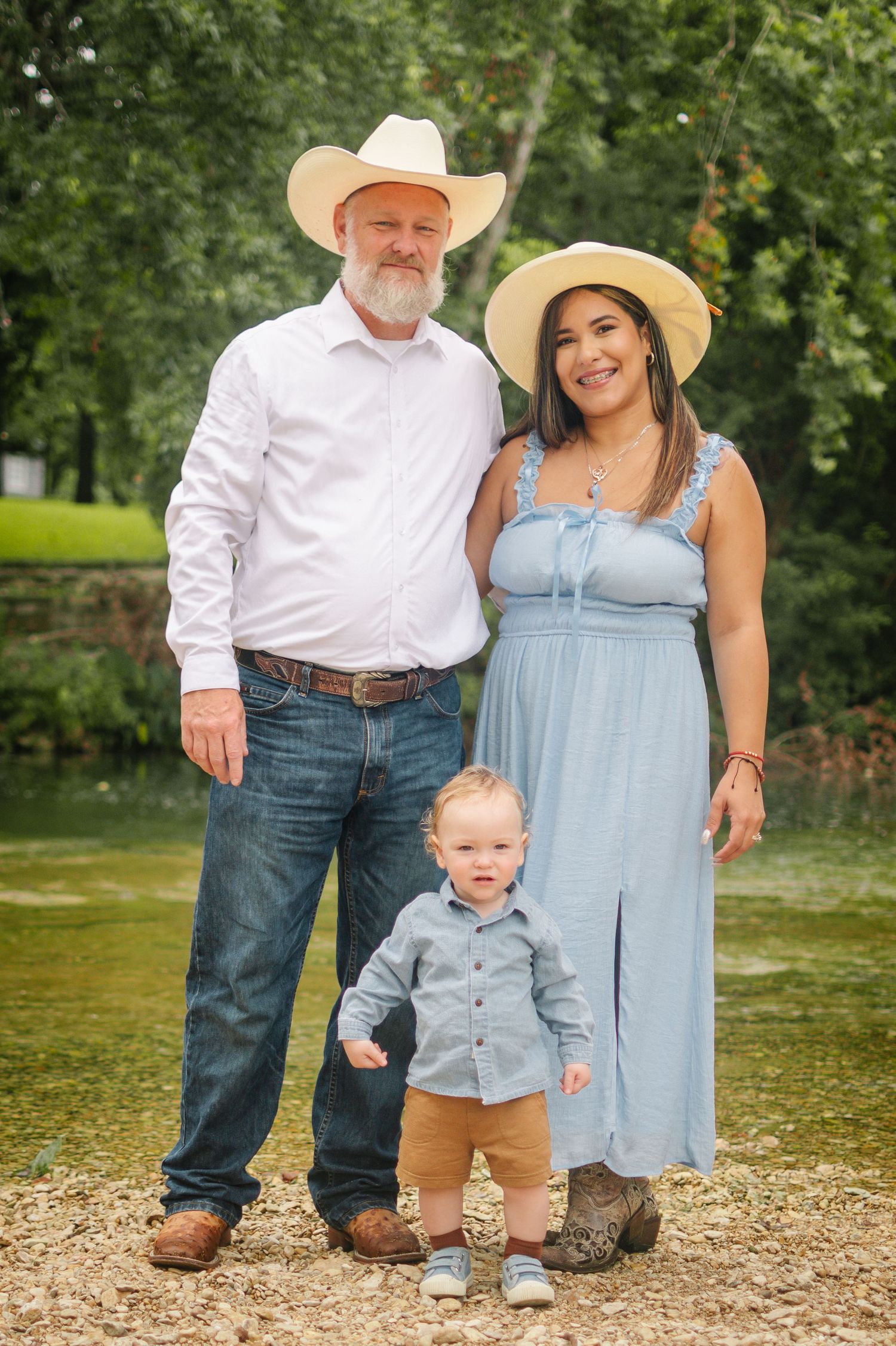A family of three poses outdoors near a creek, wearing cowboy hats and casual western attire with a toddler in front.