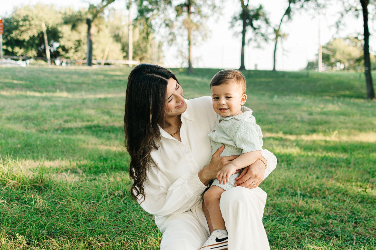 A smiling mother in white outfit holds her baby boy on her lap while sitting on green grass in a sunny park.