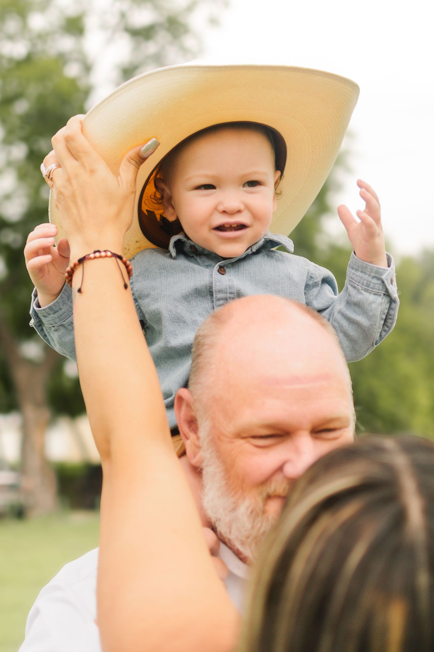 Grandparents joyfully lifting a smiling baby wearing a cowboy hat outdoors in a green park.