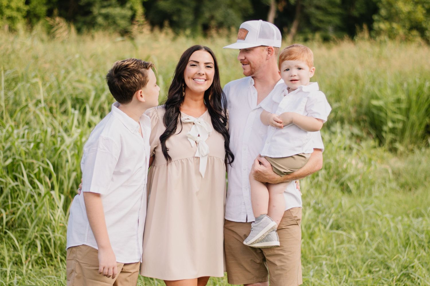 Happy family of four posing outdoors in coordinating neutral outfits, smiling together in a lush green field.