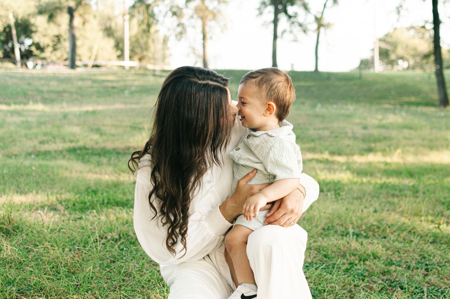 A mother holds and kisses her baby boy outdoors on green grass in a sunny park setting.