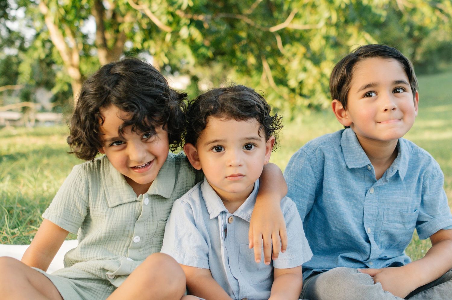 Three young brothers sit together smiling on grass in a sunny outdoor park.