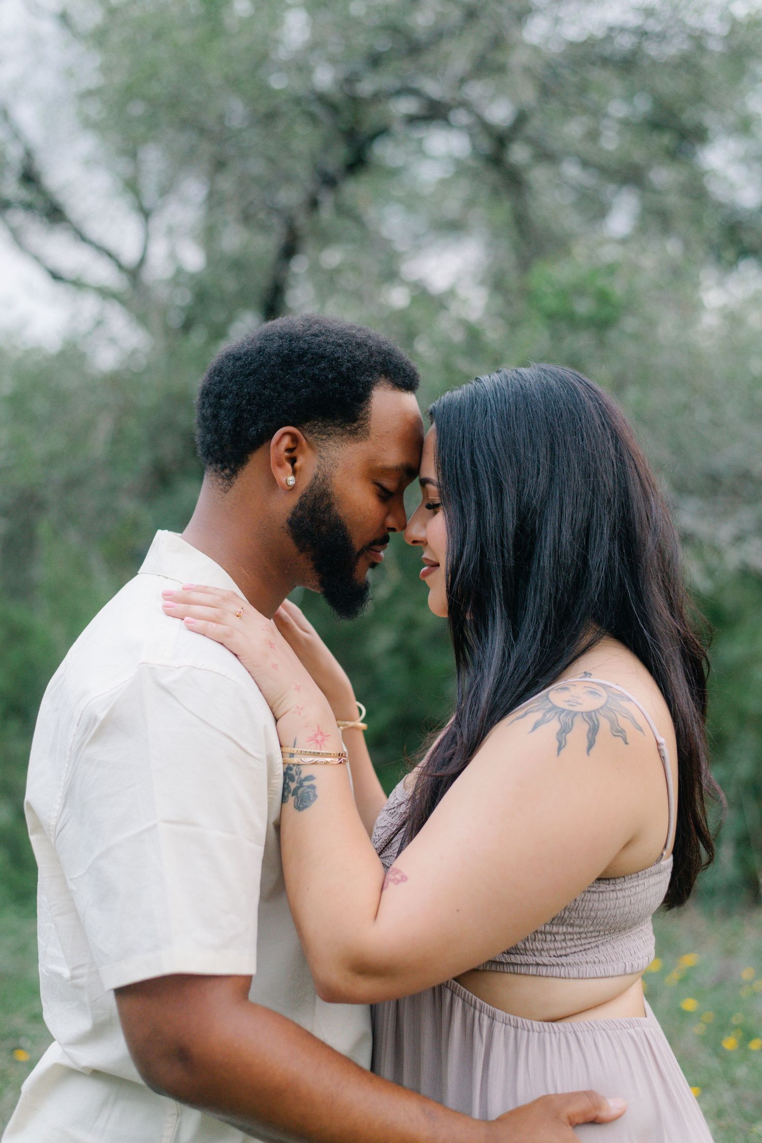A couple with tattoos shares a romantic forehead touch outdoors, surrounded by lush greenery.