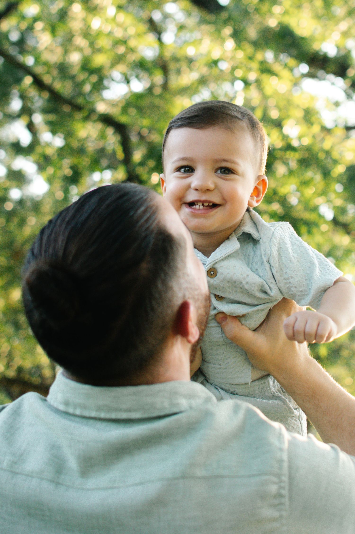 Father lifting smiling baby boy outdoors in a lush green park on a sunny day.