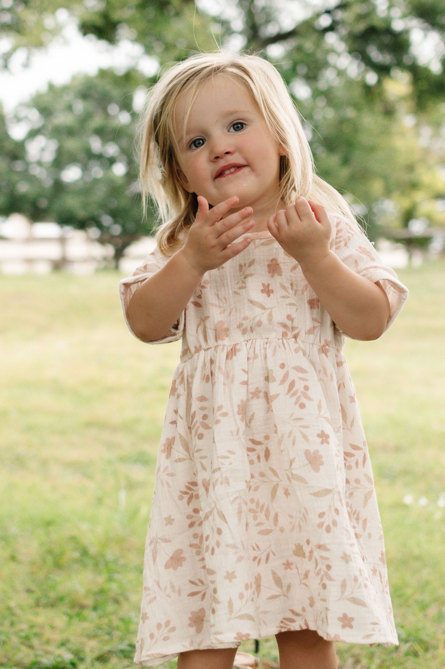Toddler girl in floral dress clapping hands outdoors in green park setting.