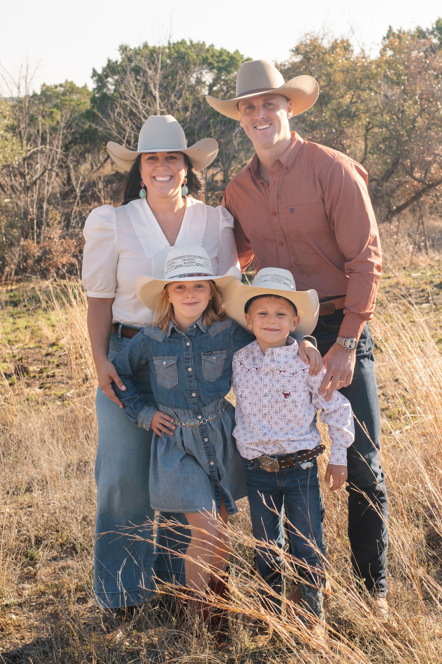 A smiling family of four in cowboy hats poses outdoors in a golden field, wearing western attire.