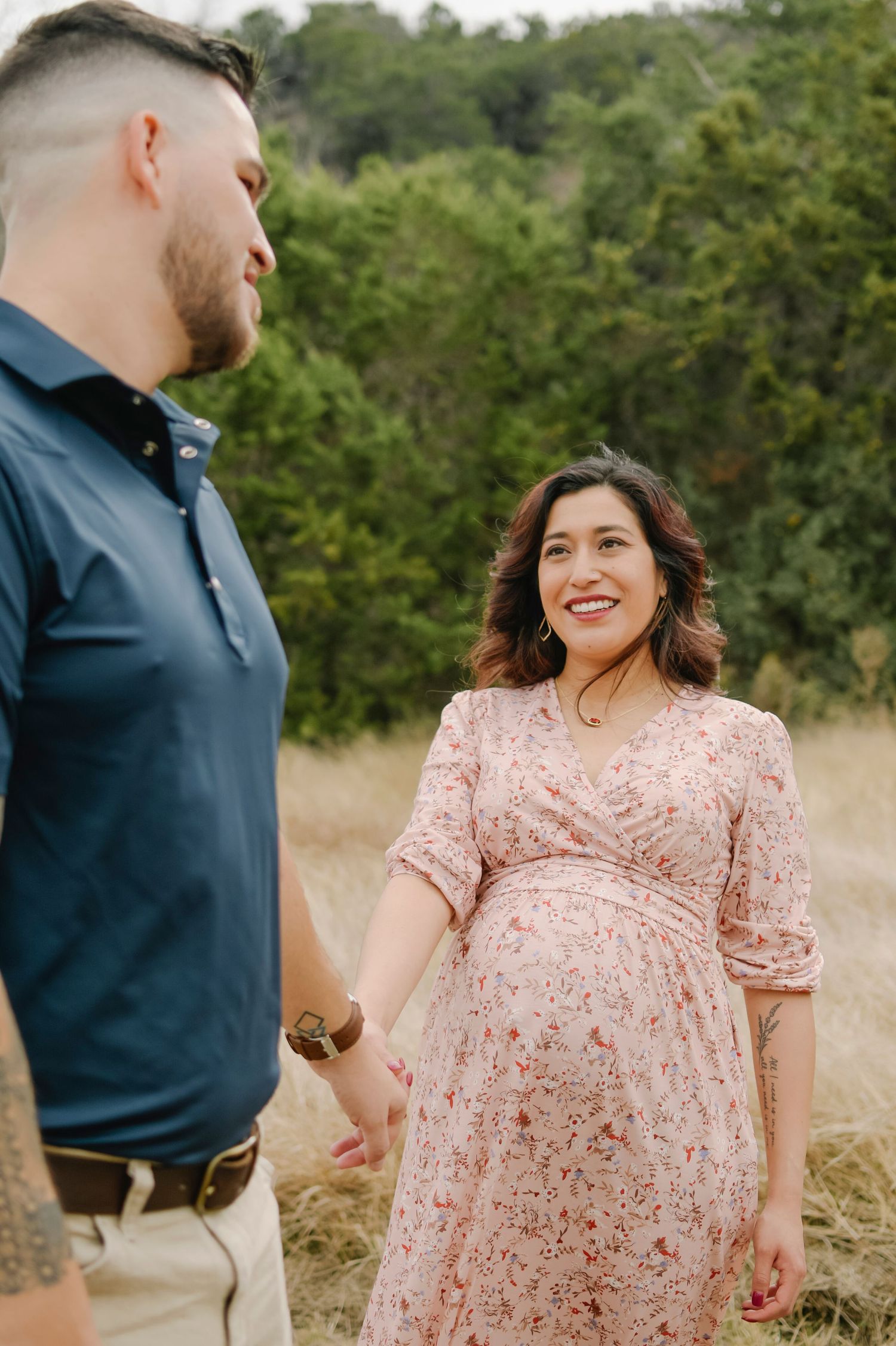 Pregnant woman in floral dress holds hands with man in navy polo in a golden field with green trees.