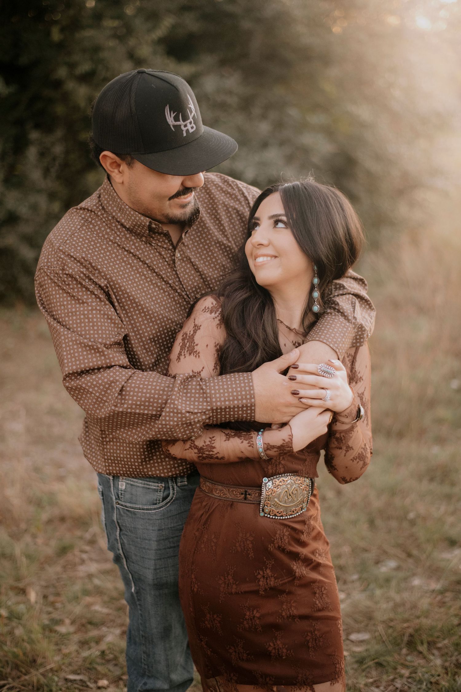Couple embracing outdoors in matching brown western outfits, woman smiling up at man wearing black snapback hat.