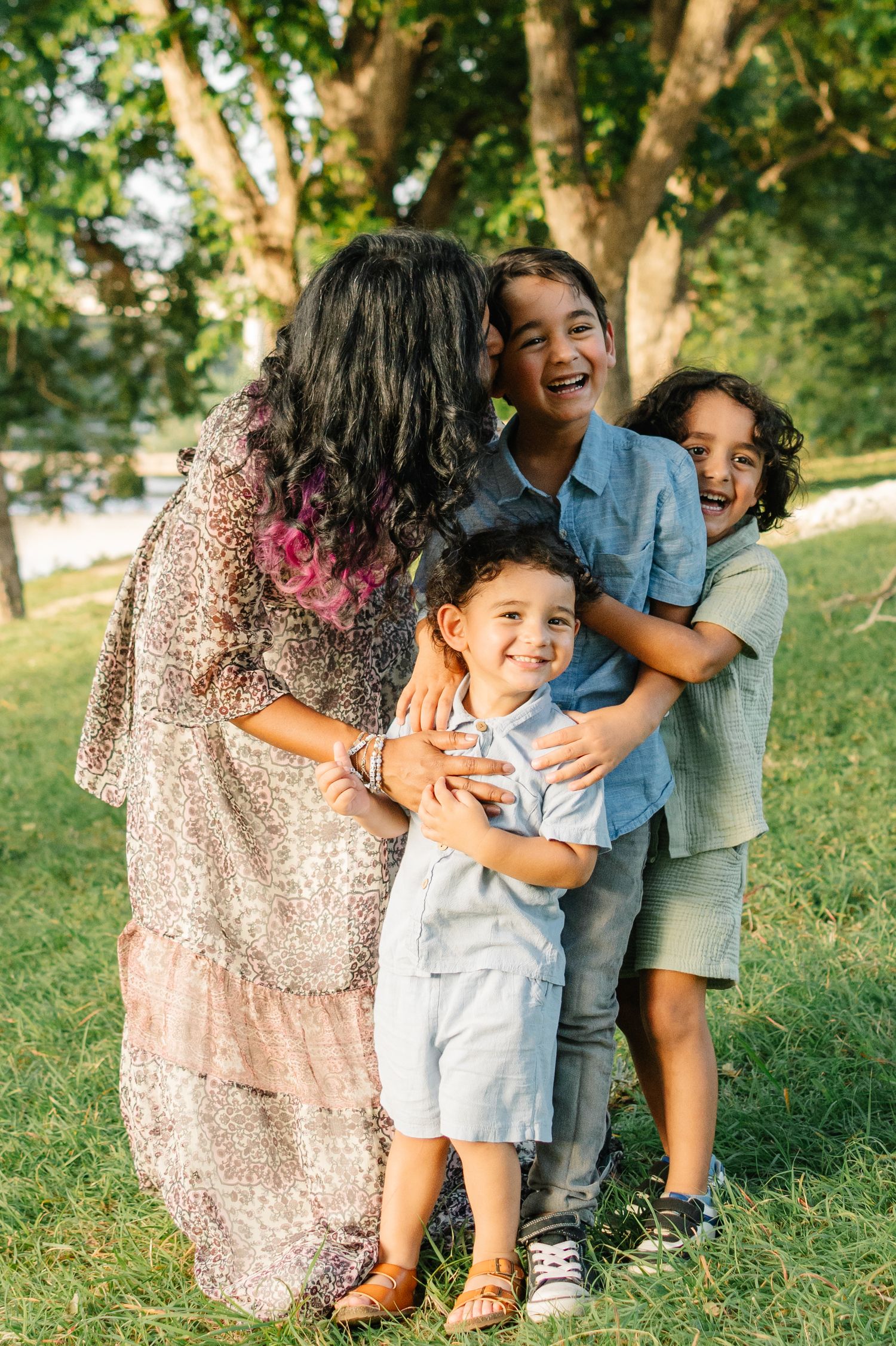 A joyful mother hugs her three laughing children outdoors in a sunny green park setting.