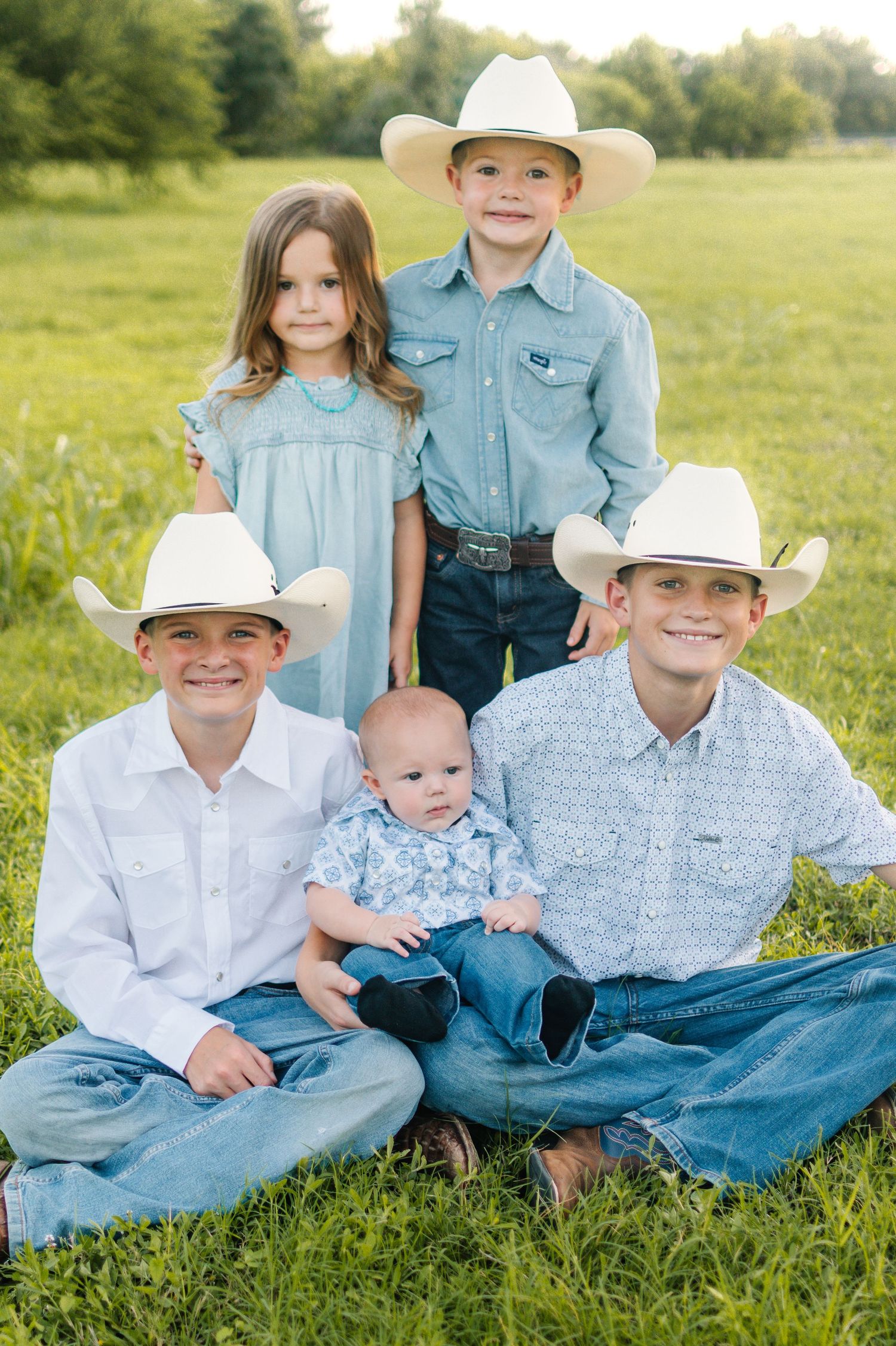 Five children in cowboy hats and denim pose together in a green field for a classic Western-style family portrait.