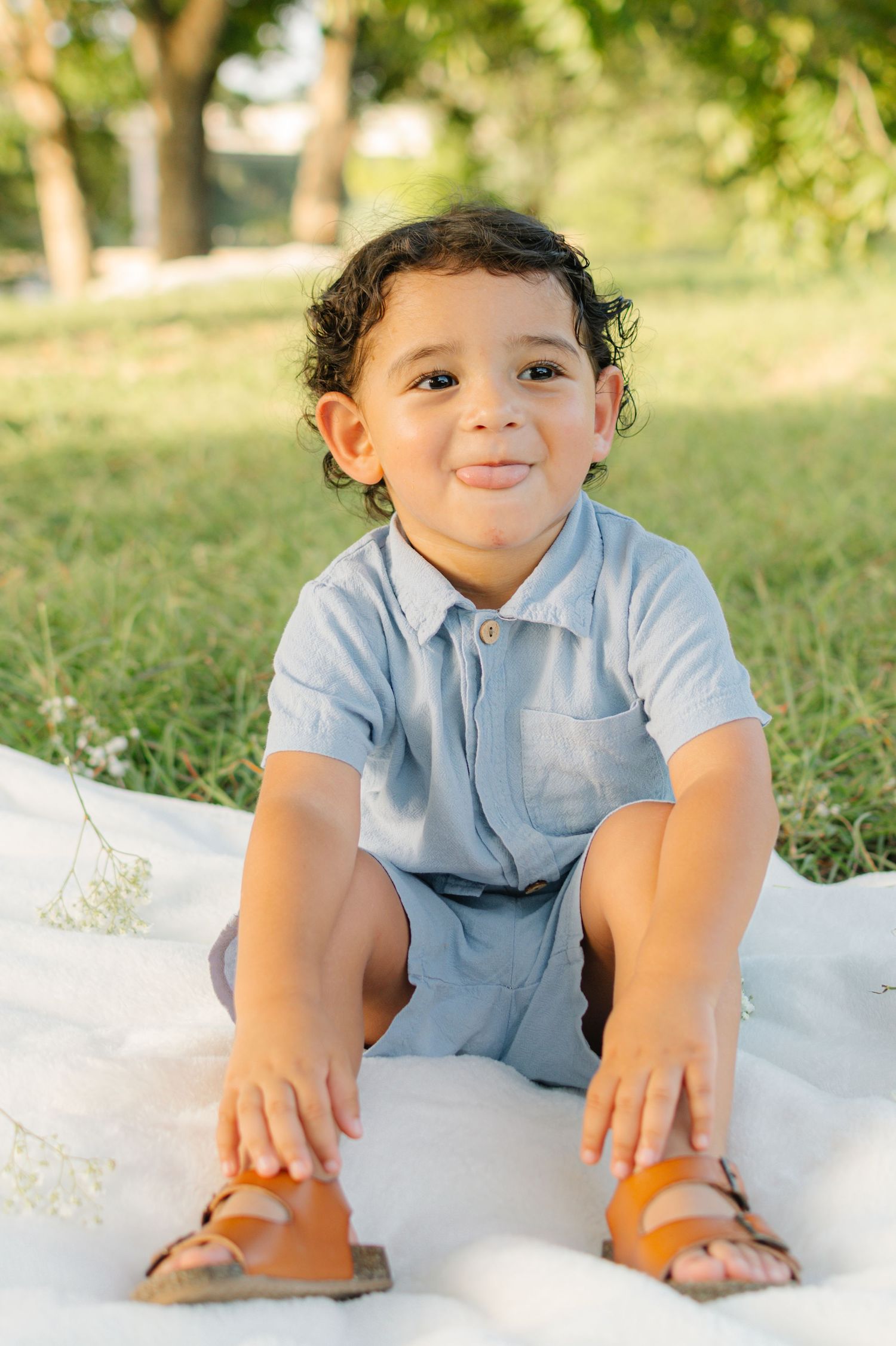 A young toddler boy with curly hair sits on a white blanket in a park, wearing a light blue outfit and brown sandals.